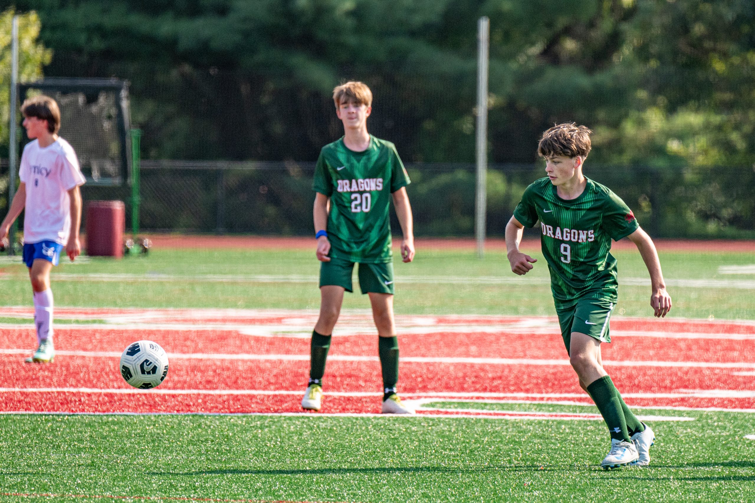 Three male student athletes playing soccer on turf field. Two boys are wearing green soccer uniforms and one boy is wearing a white soccer uniform.