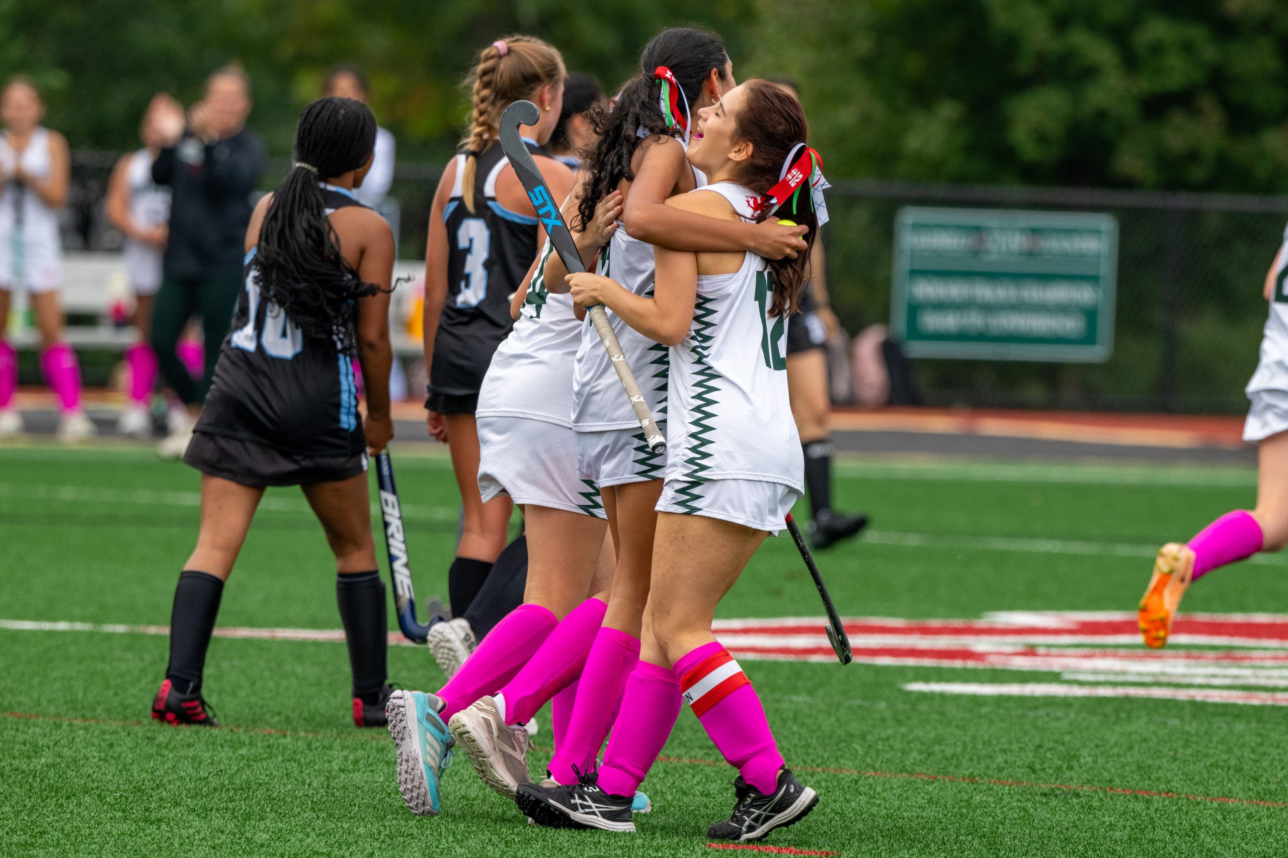Three female student athletes celebrate on turf field. The three girls are wearing white field hockey uniforms.