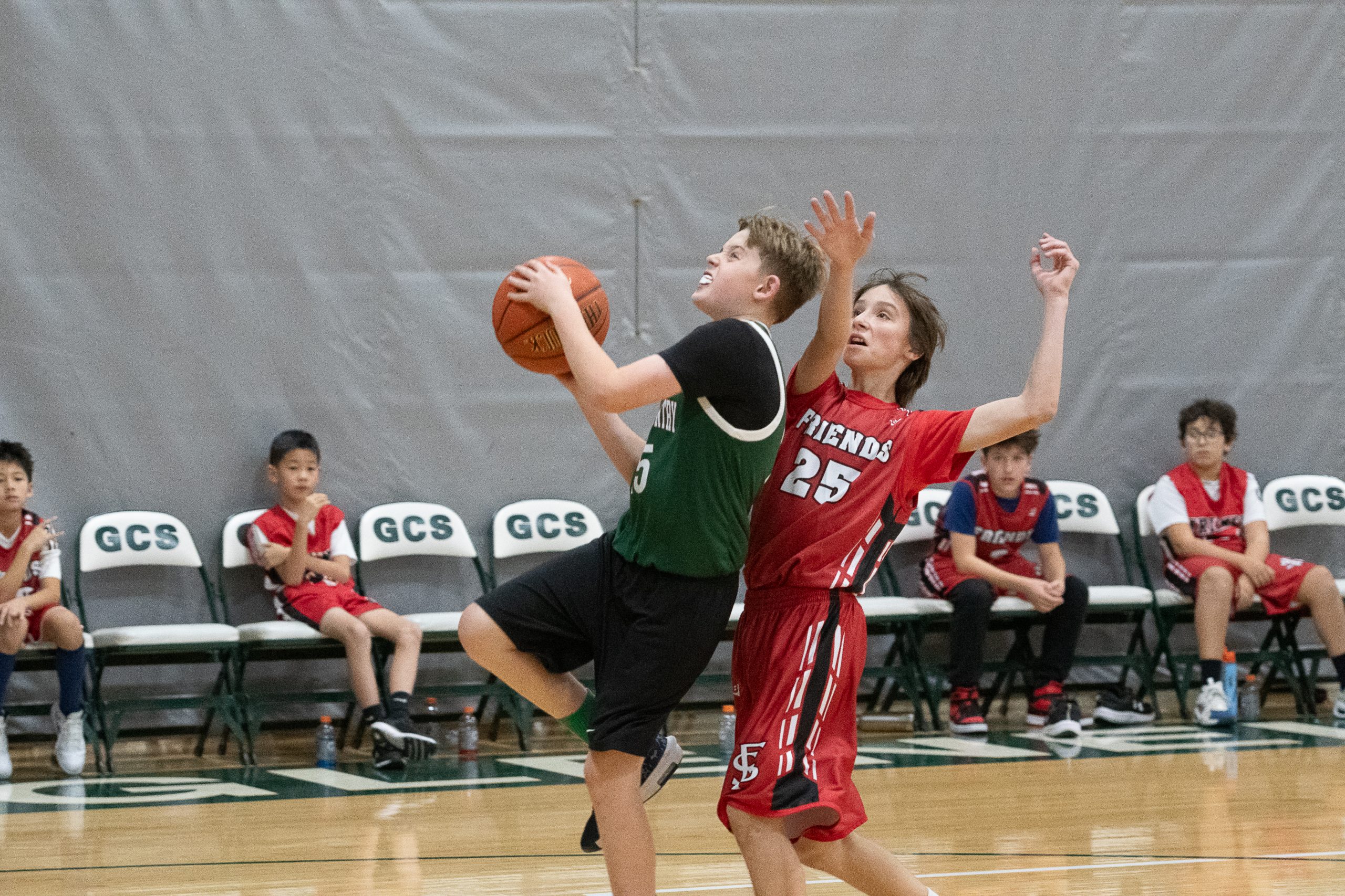 Two male athletes playing basketball on basketball court. One boy wears a green basketball uniform, and one boy wears a red basketball uniform. One boy is shooting the ball.