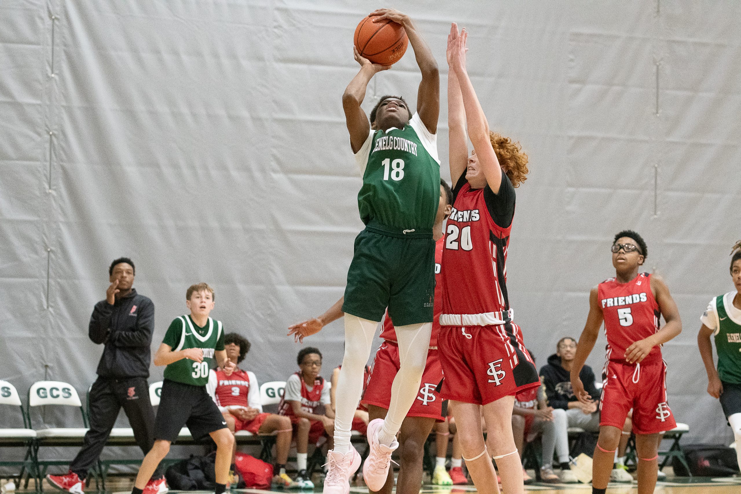 Five male student athletes play basketball on basketball court. Three boys wear a red basketball uniform, and two boys wear green basketball uniforms. One boy is shooting basketball.