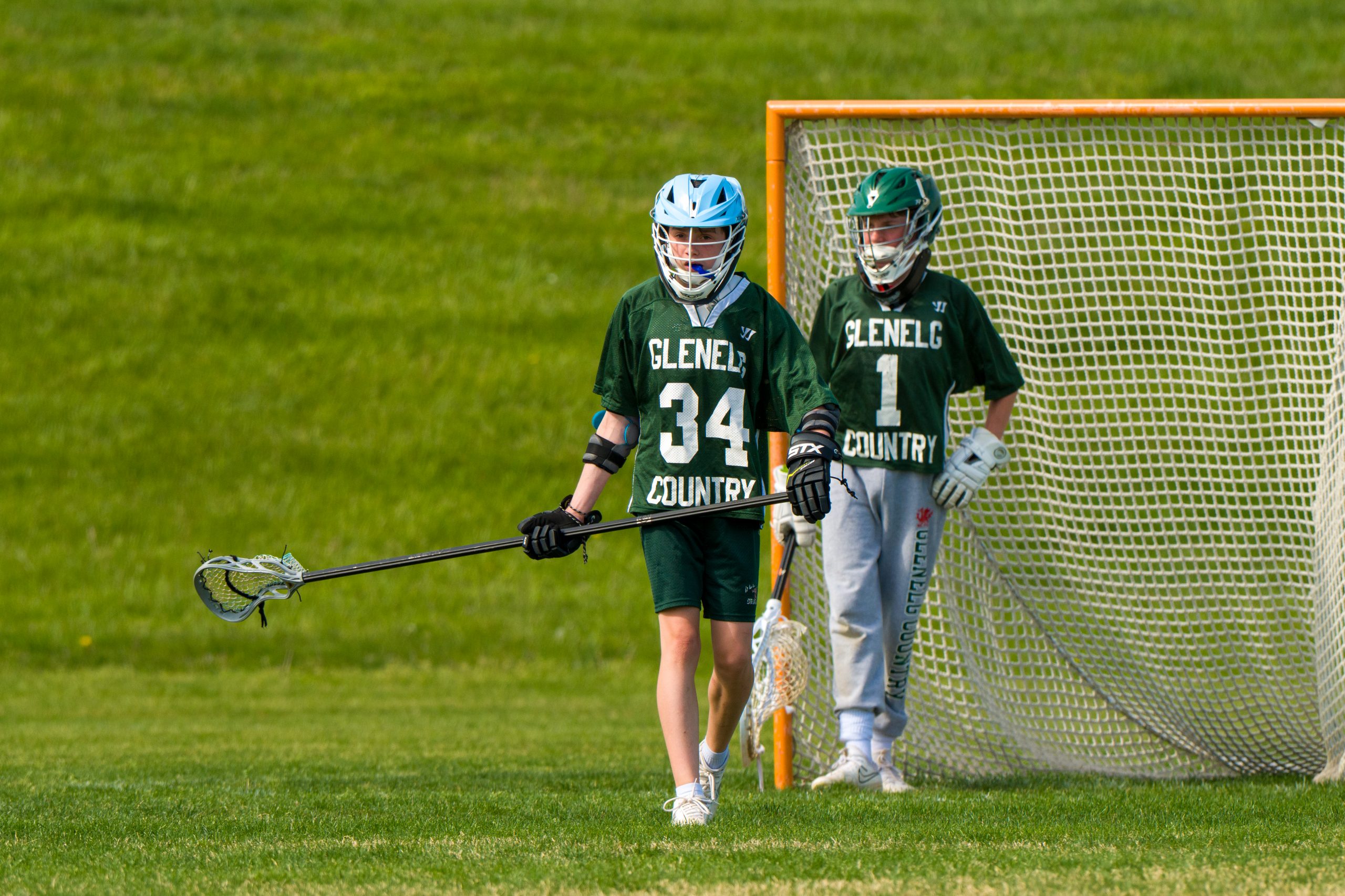 Two male student athletes play lacrosse. Both boys are wearing green lacrosse uniforms and hold lacrosse sticks.