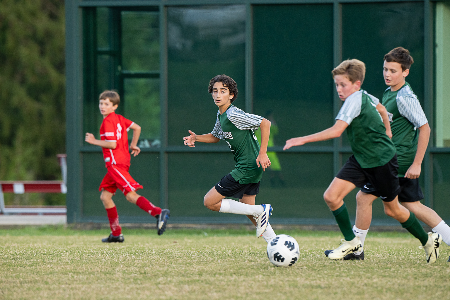 Four male student athletes playing soccer on grass field. Three boys wear green soccer uniforms, and one boy wears a red soccer uniform. One boy is about to kick the ball.