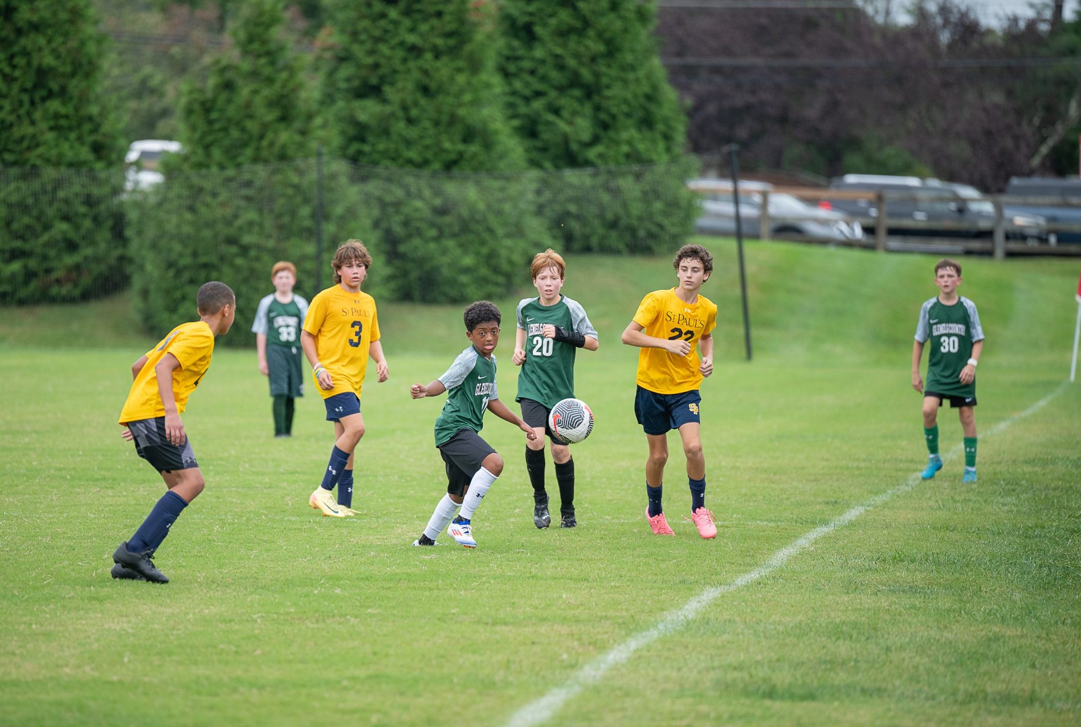 Seven male student athletes play soccer on grass field. Four boys wear green soccer uniform, and three boys wear yellow soccer uniform.