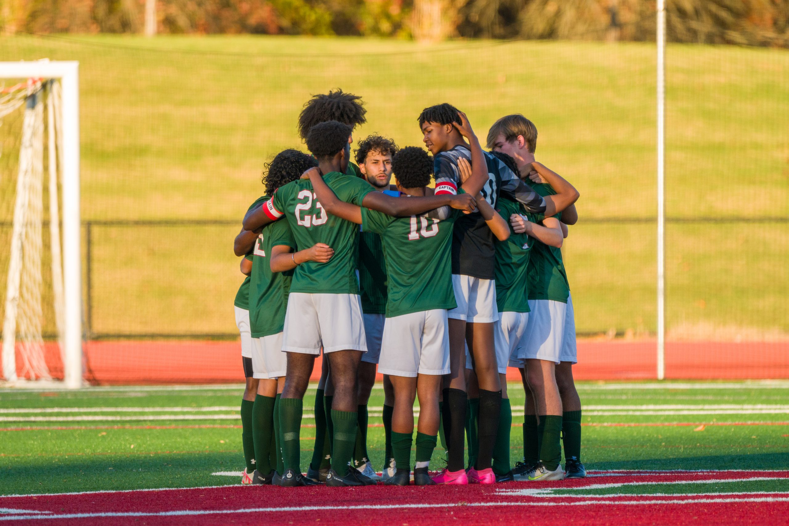 Group of 7 male student athletes on soccer field. 6 boys wearing green soccer uniform, one boy wearing black soccer jersey.