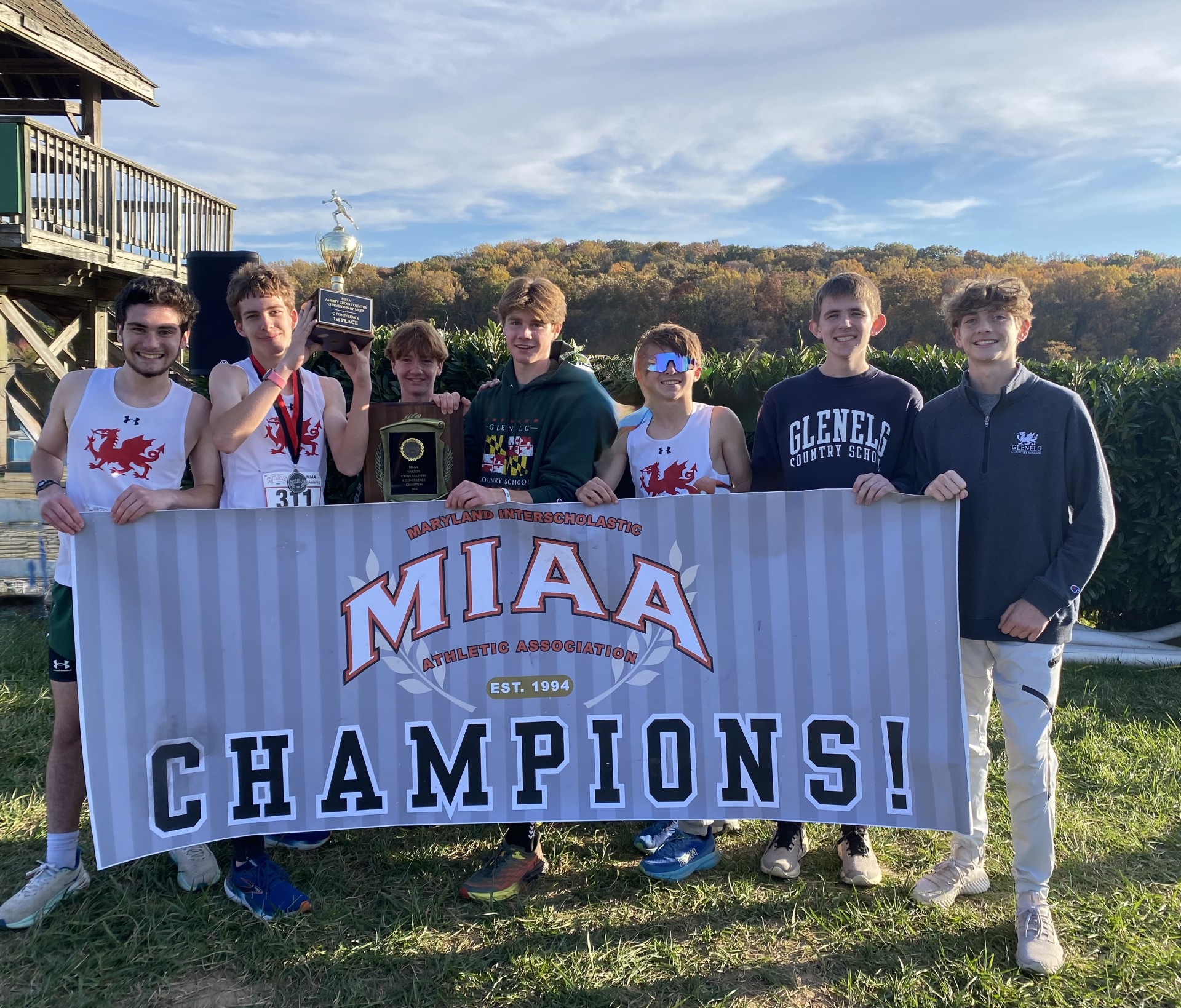 Seven students stand holding a championship banner. Three boys wear white sleeveless shirts. Two boys wear a grey long sleeved shirt. One boy wears a green long sleeve sweatshirt.