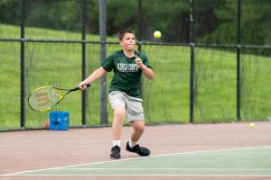Male student athlete playing tennis on tennis court. Boy wears green short sleeve shirt and silver shorts.