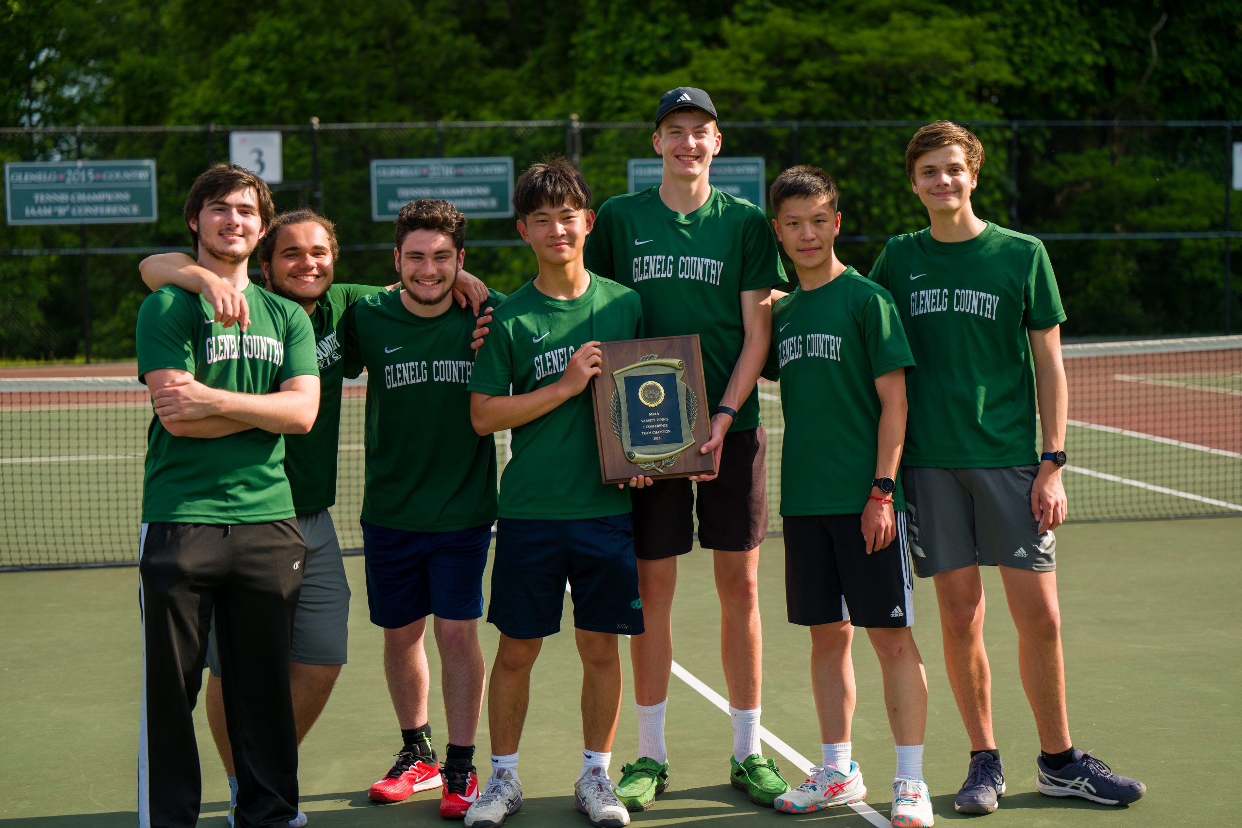 Seven male student athletes wearing green tennis uniforms holding trophy on tennis courts.