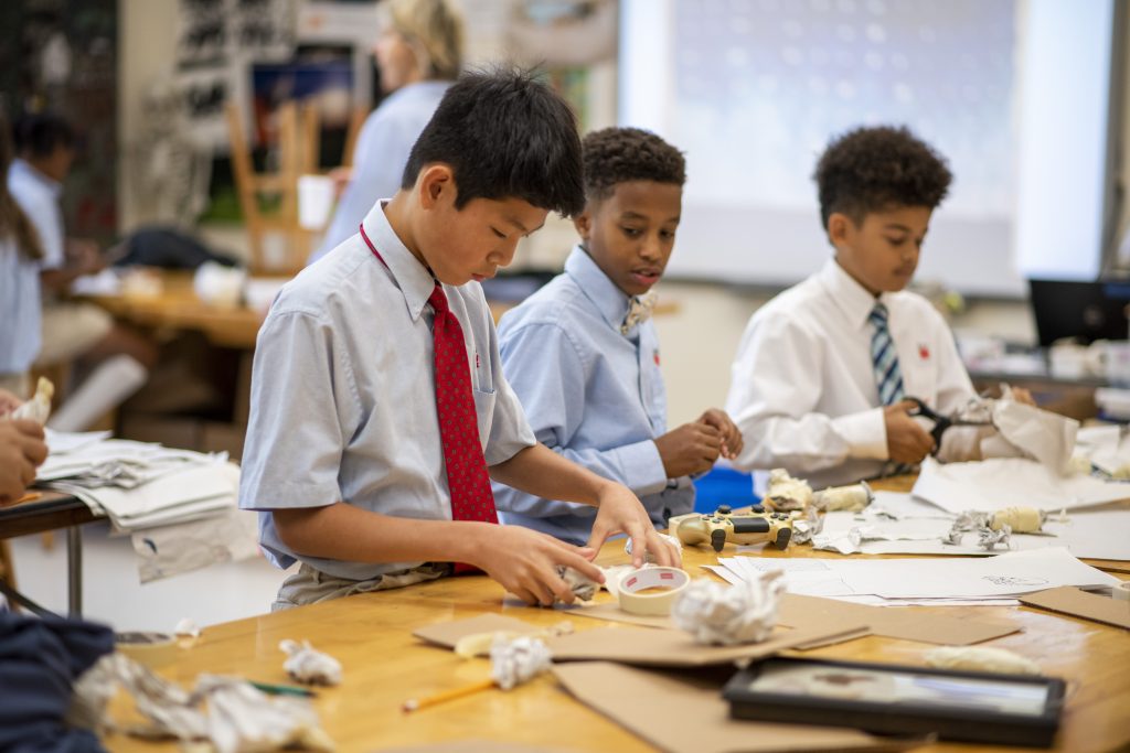 Three boys work on an art project at a long table.