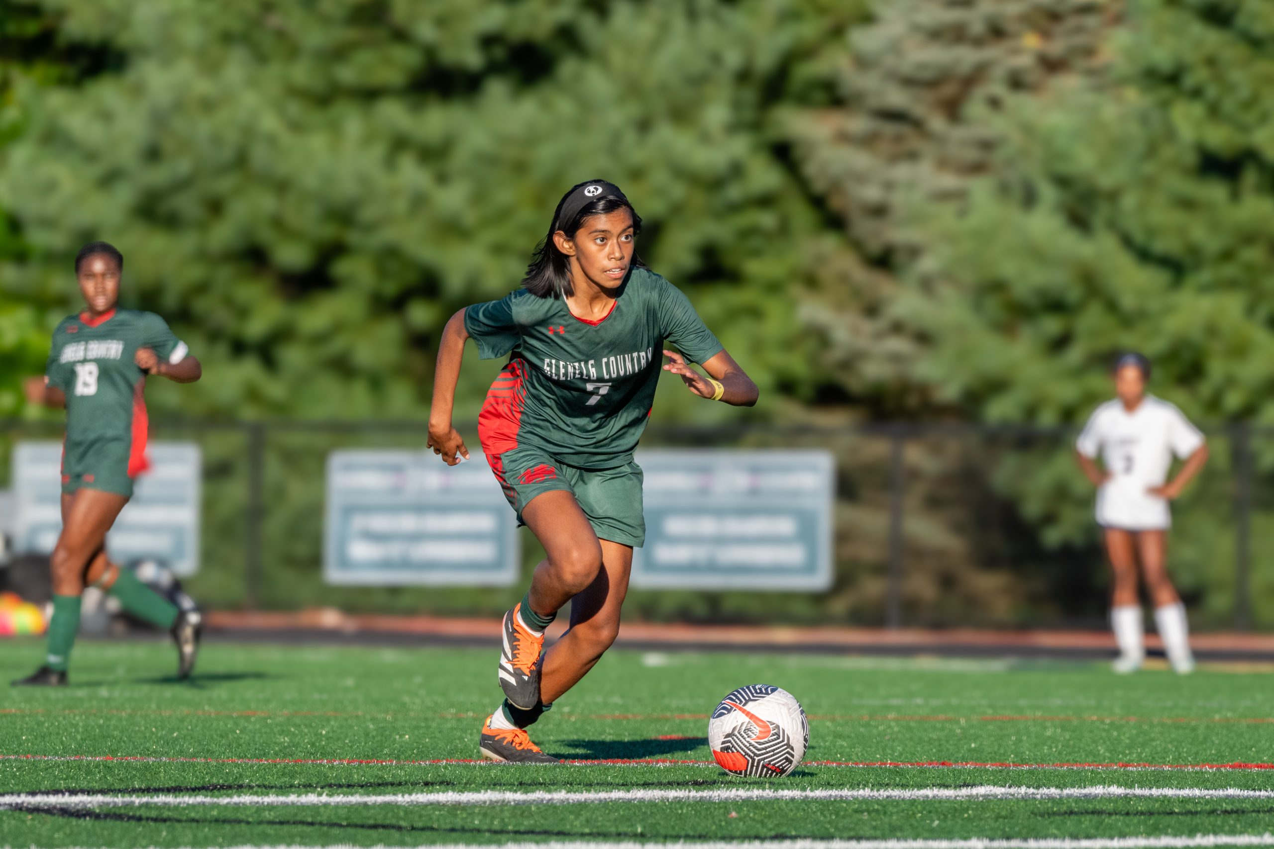 Female student athlete will soccer ball on soccer field, wearing green soccer uniform.