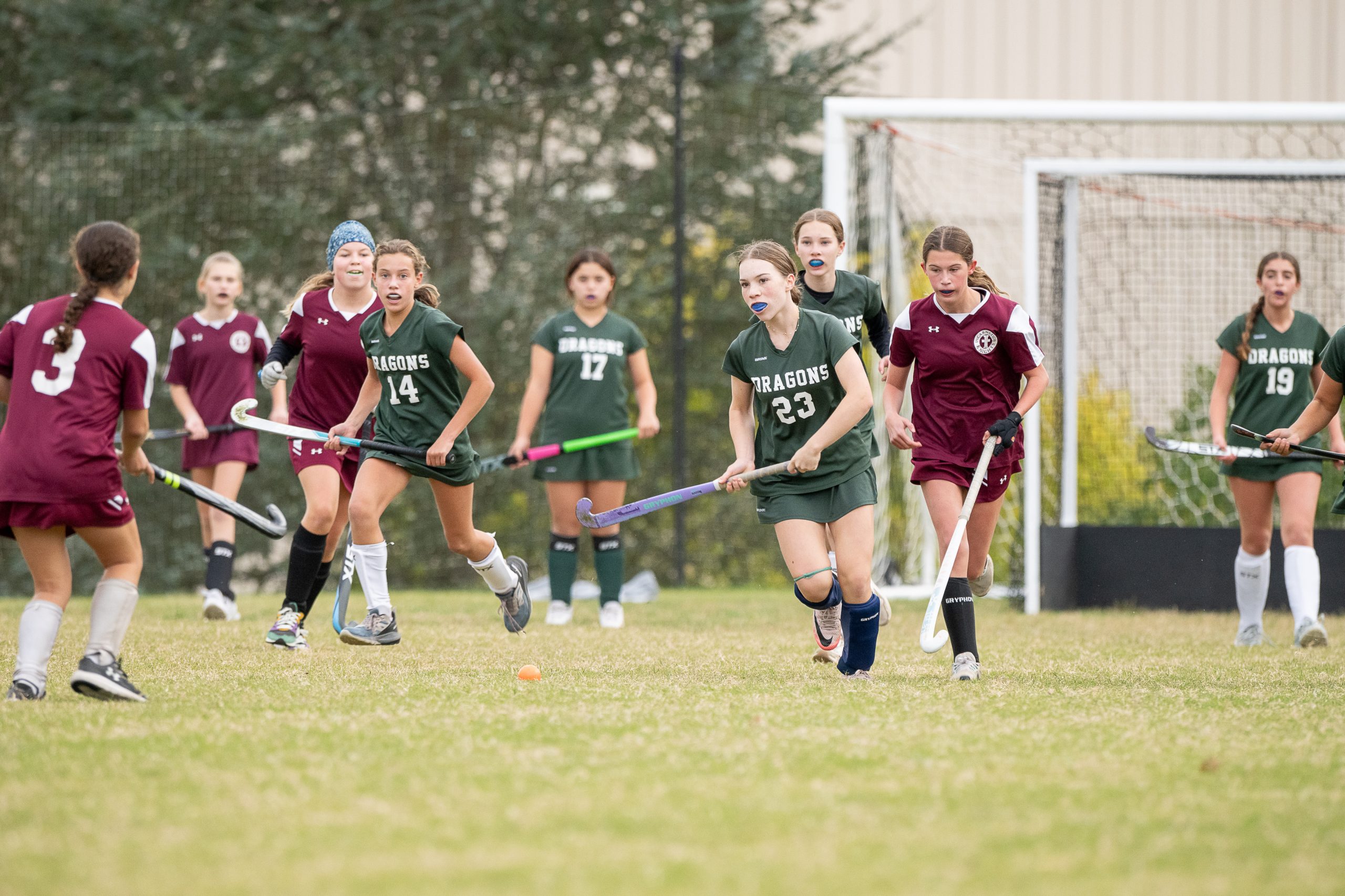 Nine female student athletes play field hockey on a grass field. Five girls wear green field hockey uniforms and four girls wear red field hockey uniforms.