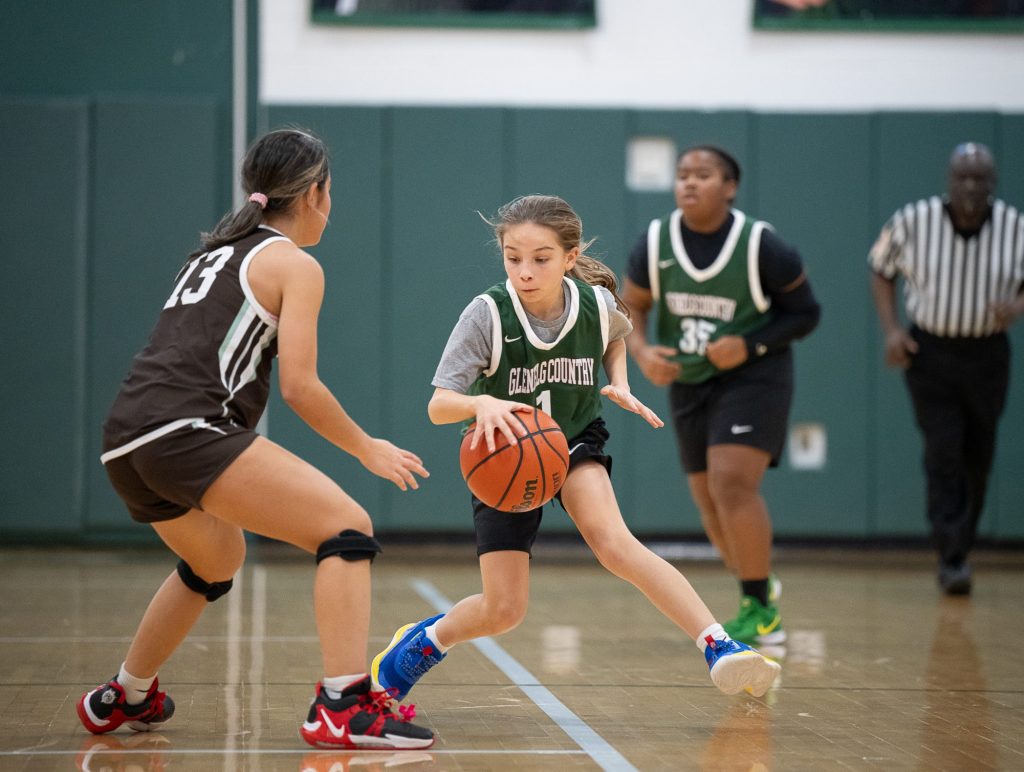 Three female student athletes playing basketball. Two girls wear green basketball uniformes, while one girl wears a brown basketball uniform. One male official in short sleeve black and white stripped top and black pants.