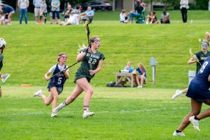 Two female student athletes play lacrosse. One wears a green lacrosse uniform, and one wears a blue lacrosse uniform. Both hold lacrosse sticks.