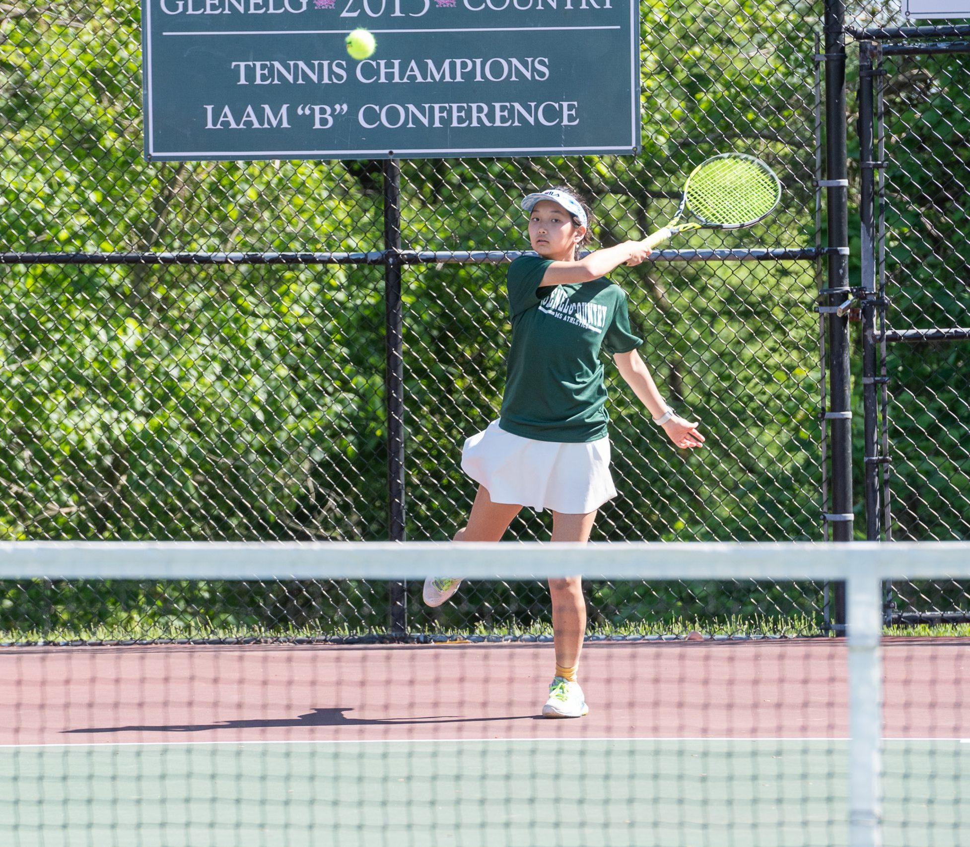 Female student athlete wearing green short sleeve top and a white skirt plays tennis on tennis courts.