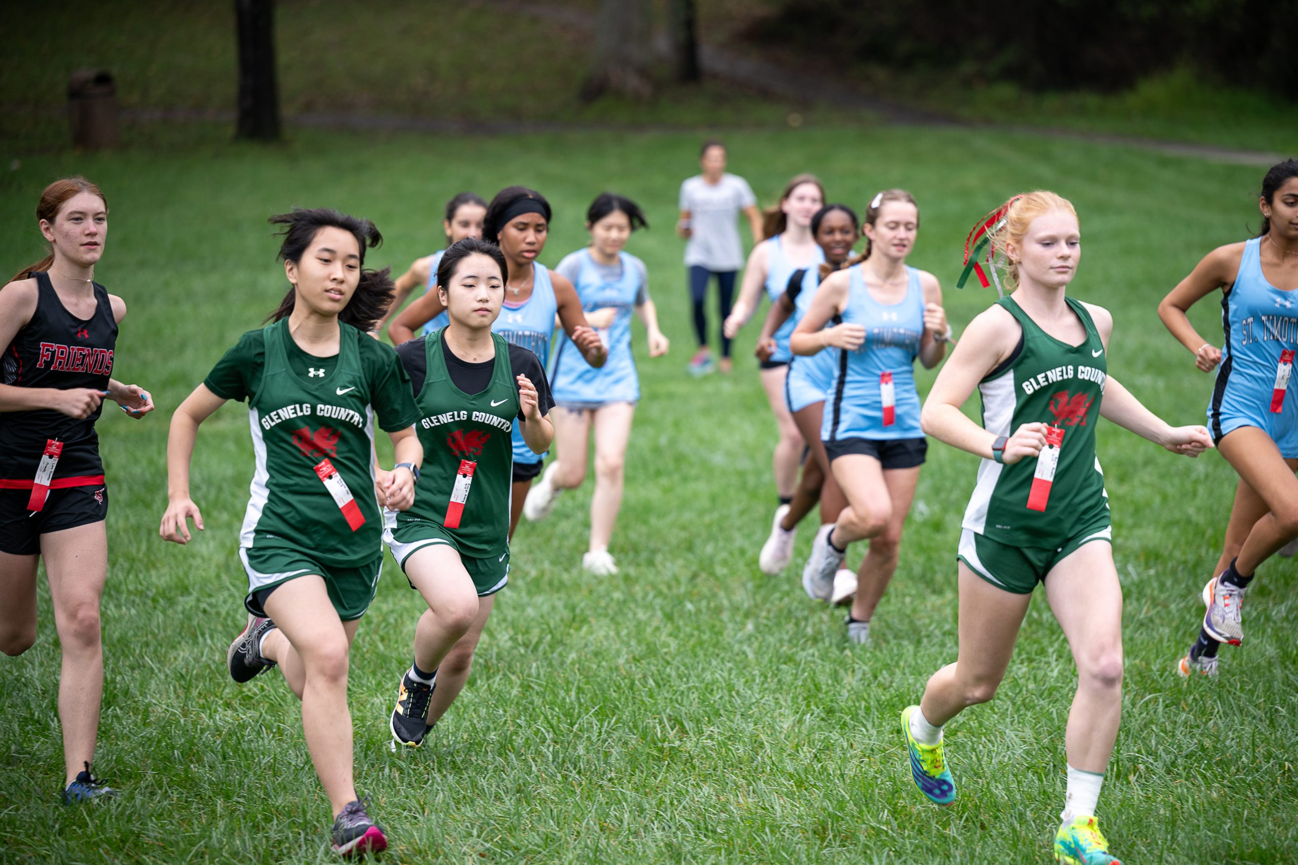 Group of 8 female student athletes running. Four females wear a blue cross-country uniform, three female athletes wear a green cross-country uniform, and one female wears a brown cross-country uniform.