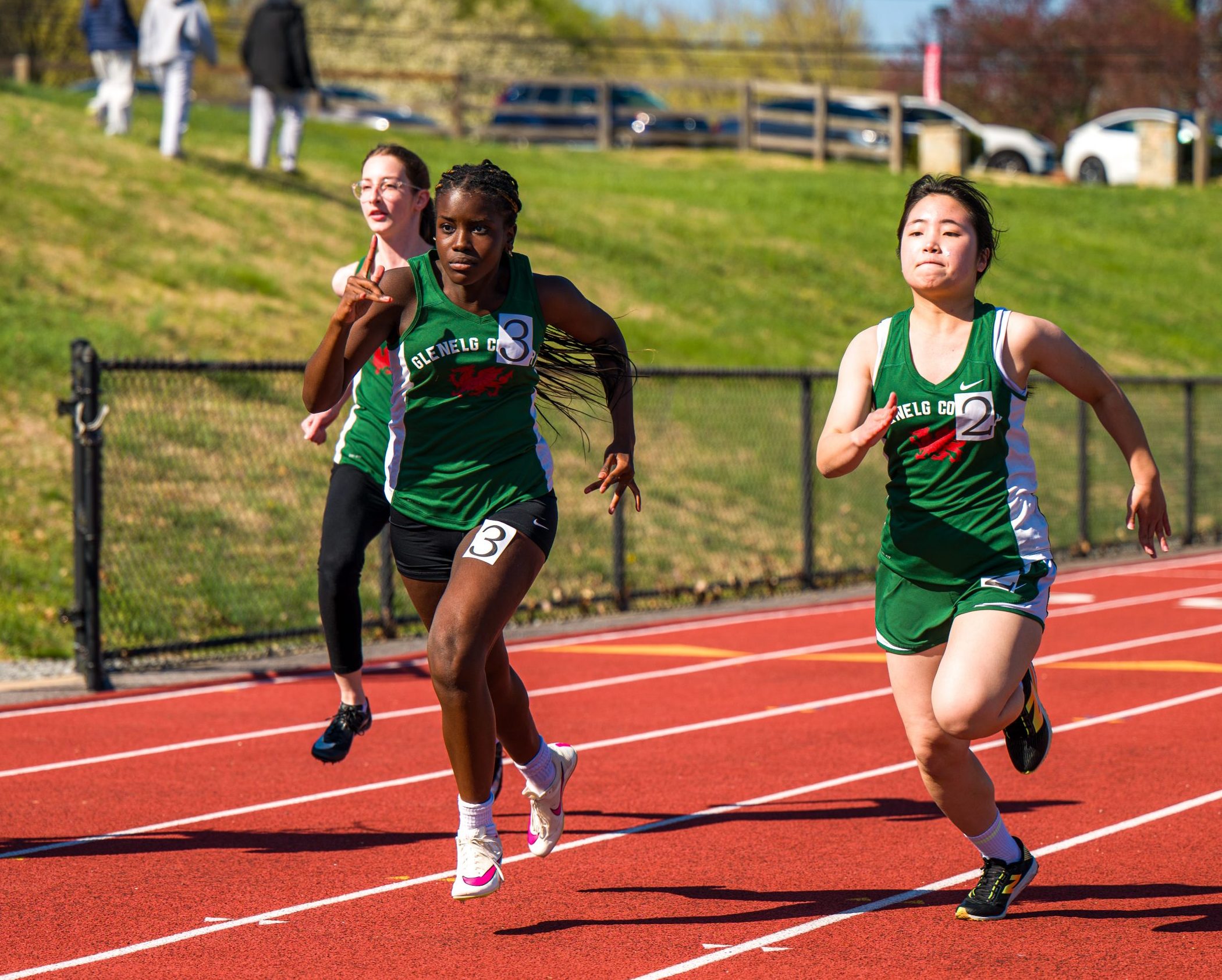 Three female student athletes run on a track. All three girls are wearing green track uniforms.