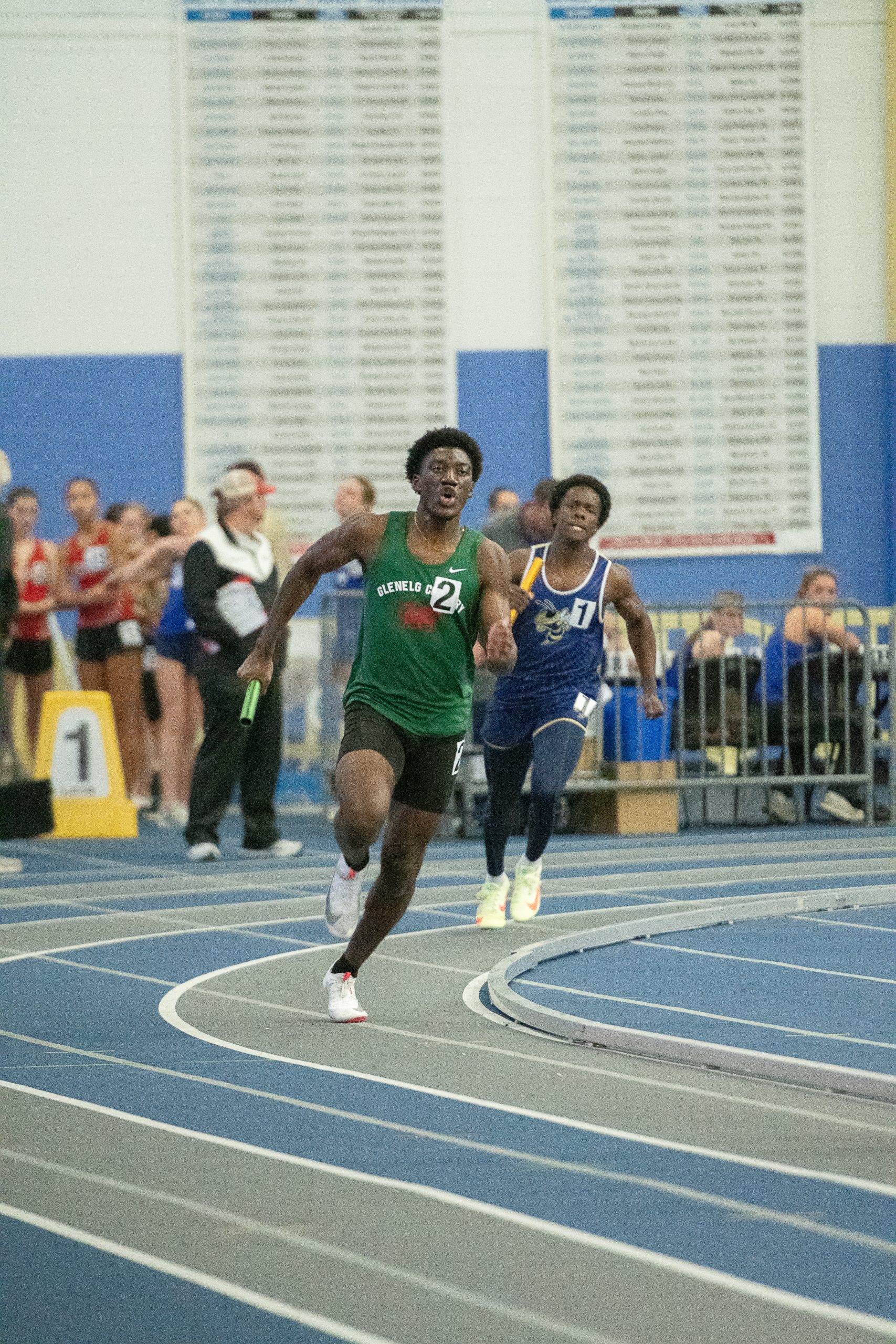 Two male athletes running track. One boy wears a green track uniform and one boy wears a blue track uniform.