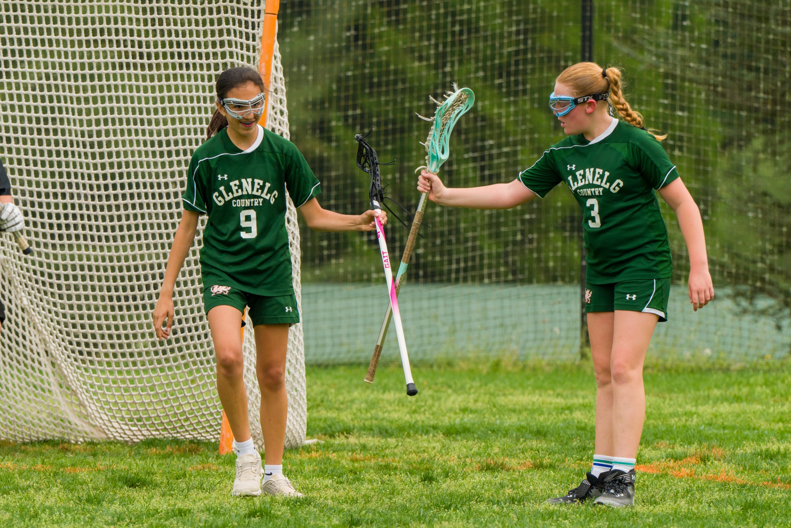Two female student athletes in green lacrosse uniforms hitting sticks together on lacrosse field.