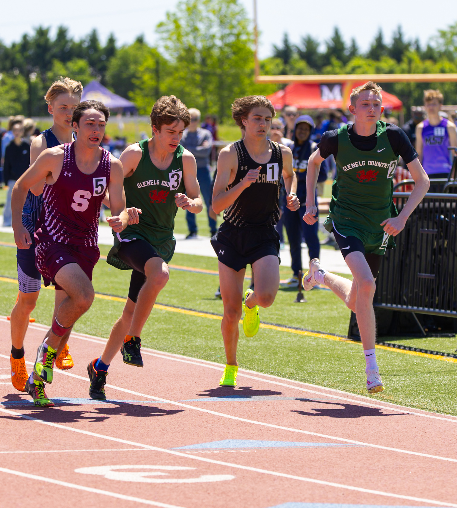 Four male student athletes running on a track. Two boys wearing a green track uniform, one boy wearing a black track uniform, and one boy wearing a red track uniform.