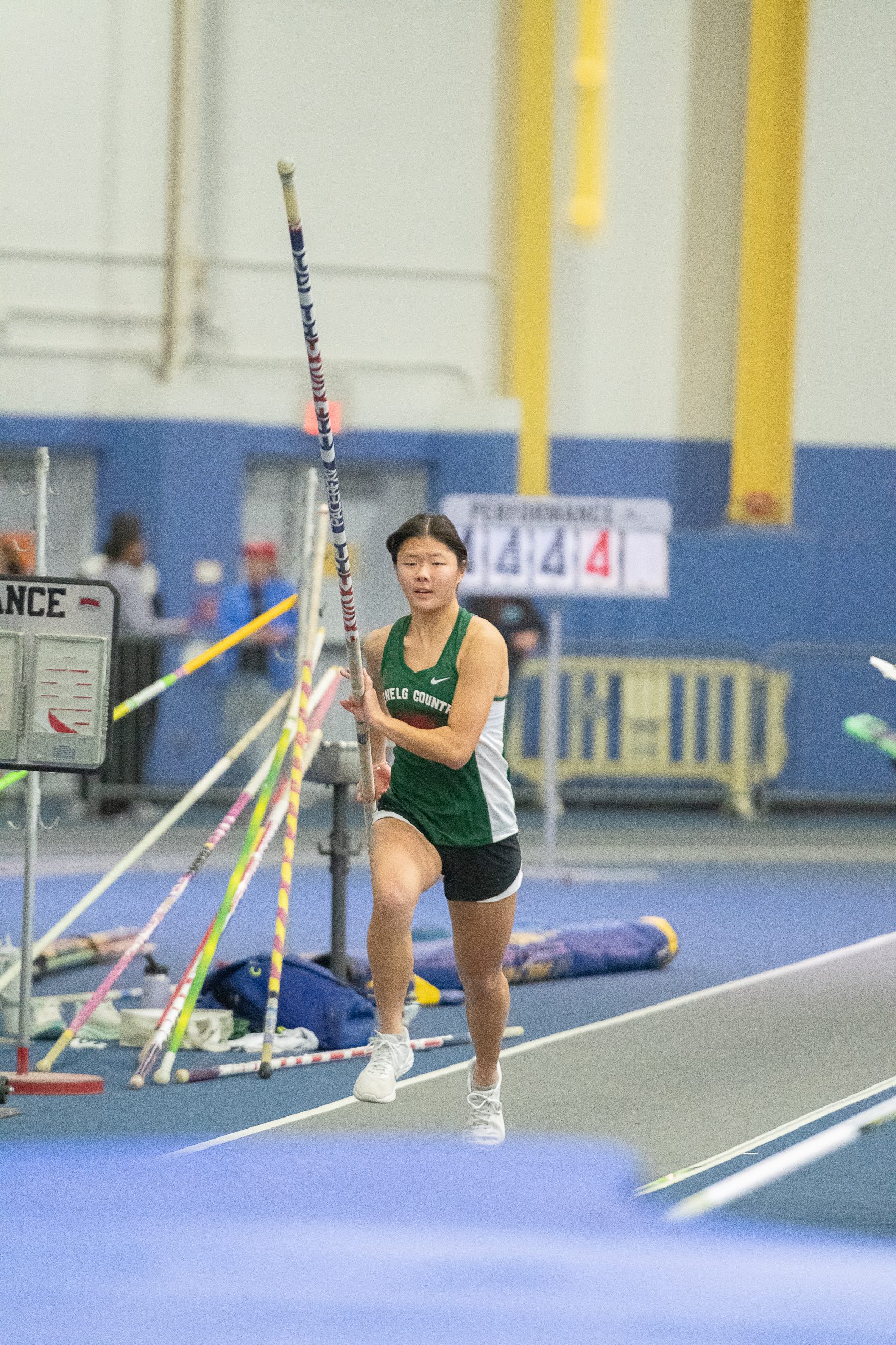Female indoor track athlete running with a pole vault pole in her hands. Athlete wears a green track uniform.