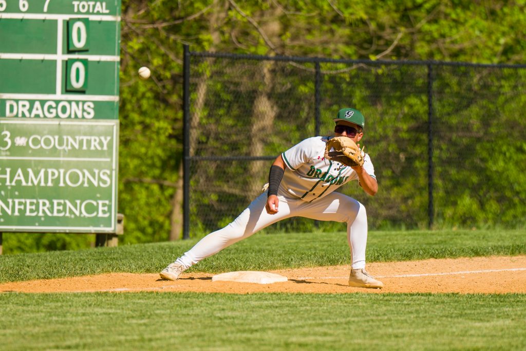 Male student athlete wearing a white baseball uniform catches ball on baseball field.