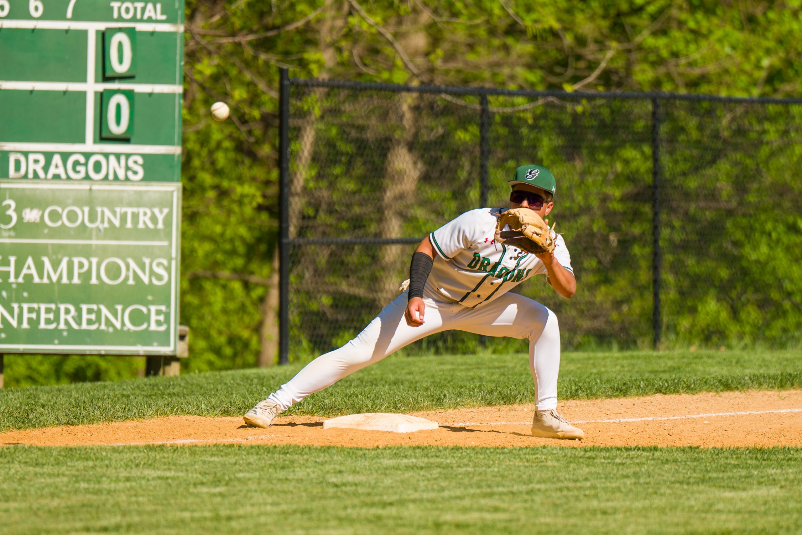Male student athlete wearing a white baseball uniform catches ball on baseball field.