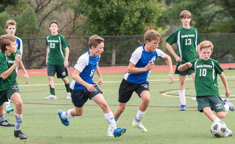 Seven male student athletes play soccer. Five students wear green uniforms and two students wear blue uniforms. One student kicks soccer ball.