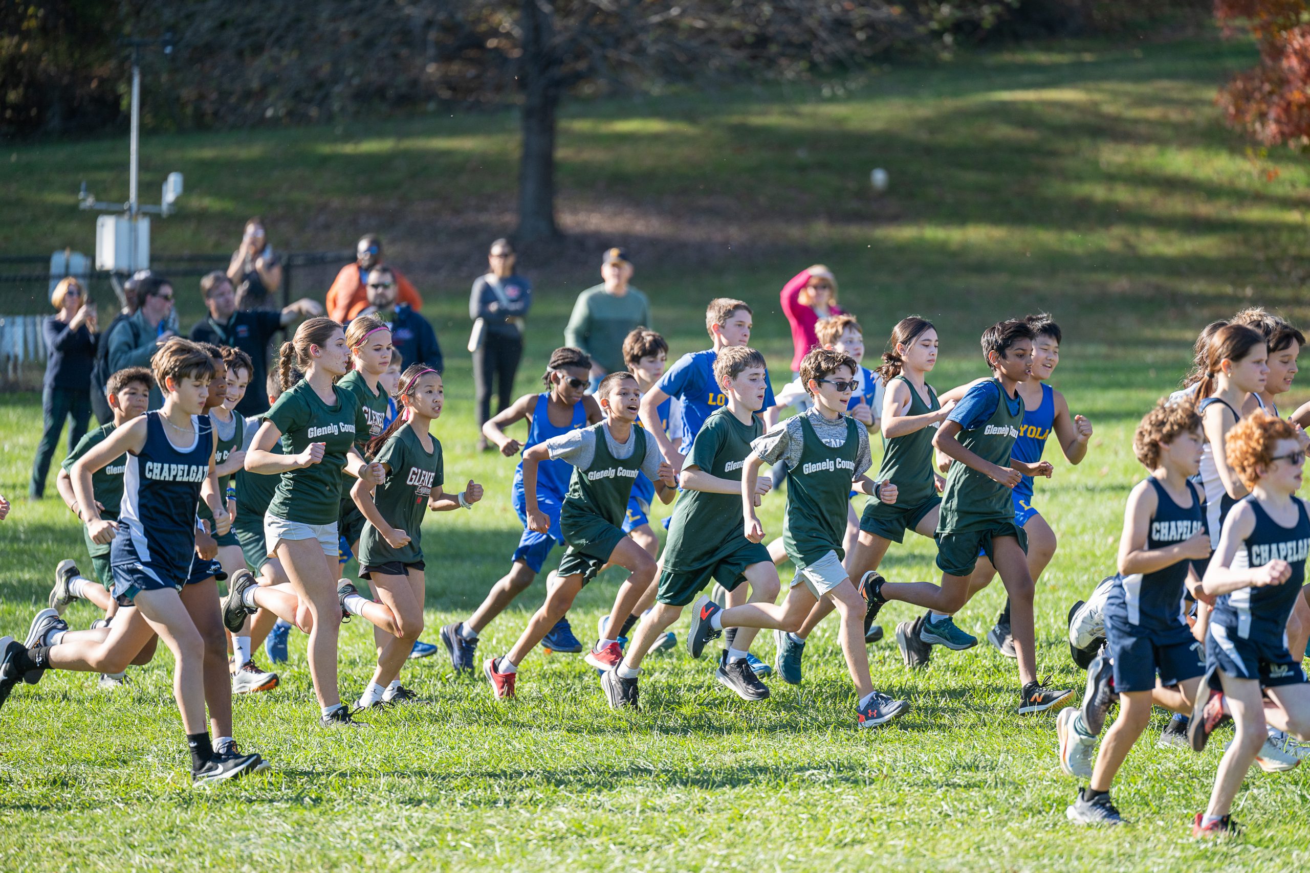 Twenty two male and female student athletes running in grass field. Eleven runners wear a blue sleeveless uniform, and nine runners wear a green sleeveless uniform.