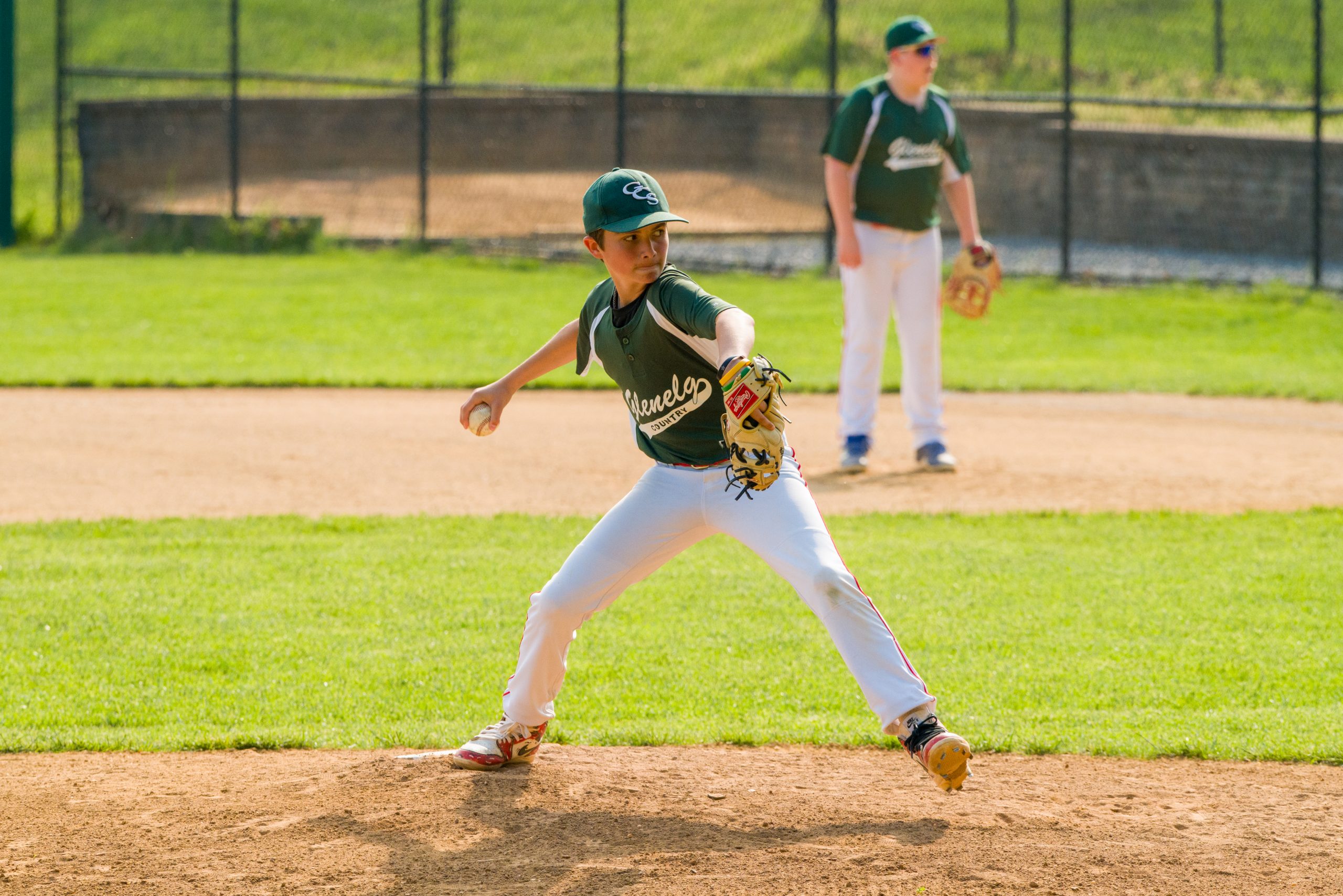 Two male athletes play baseball on baseball field. Two boys wear green baseball tops with grey baseball pants. One boy is pitching the ball.