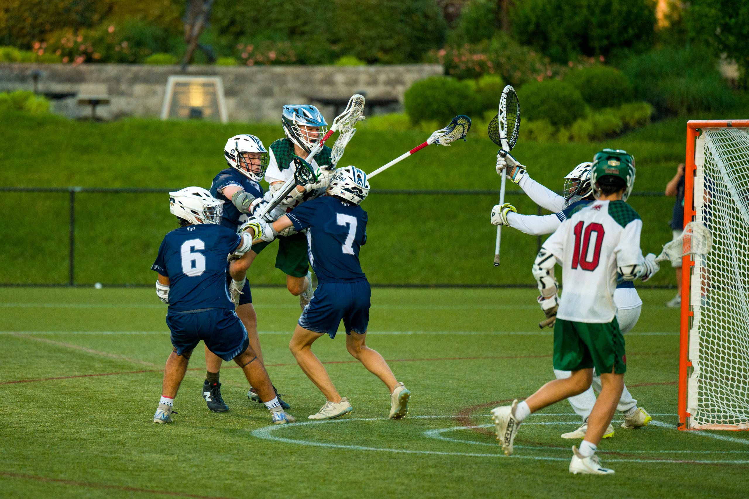Six student athletes playing lacrosse on field. Four boys wear blue lacrosse uniforms and two boys wear white lacrosse uniforms. Six boys hold lacrosse sticks.