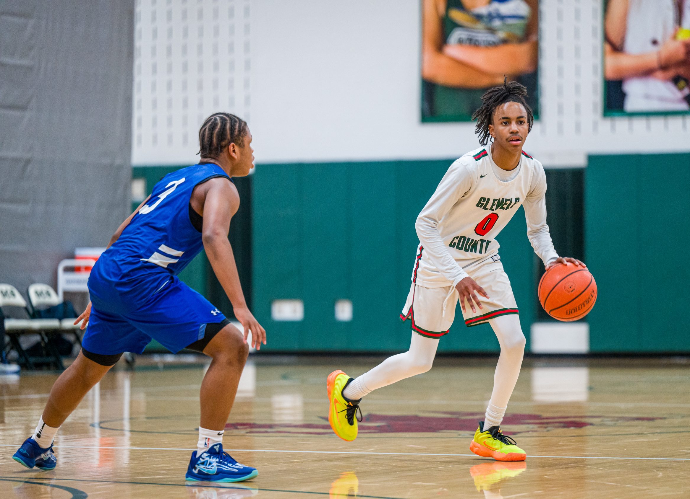 Two male student athletes play basketball on basketball court. One boy wears a white basketball uniform, and one boy wears a blue basketball uniform. One boy dribbles ball.