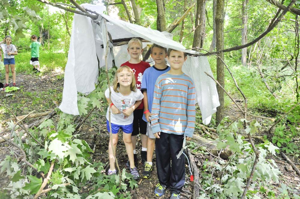 Summer in the Country campers stand underneath a tarp that is draped over a tree branch in the forest.