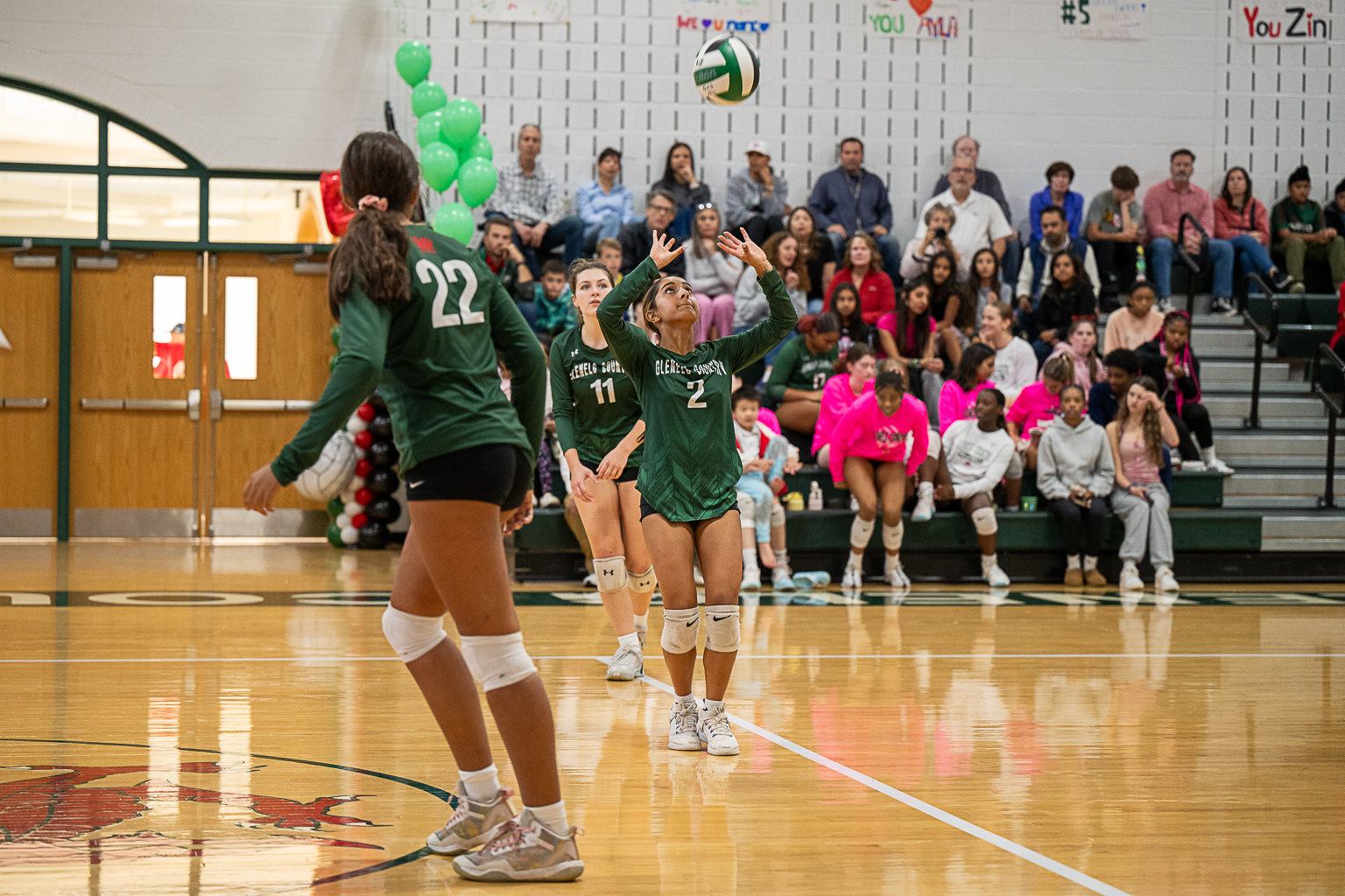 Three female student athletes play volleyball. Three girls wear green volleyball uniforms. One girl is setting the ball.