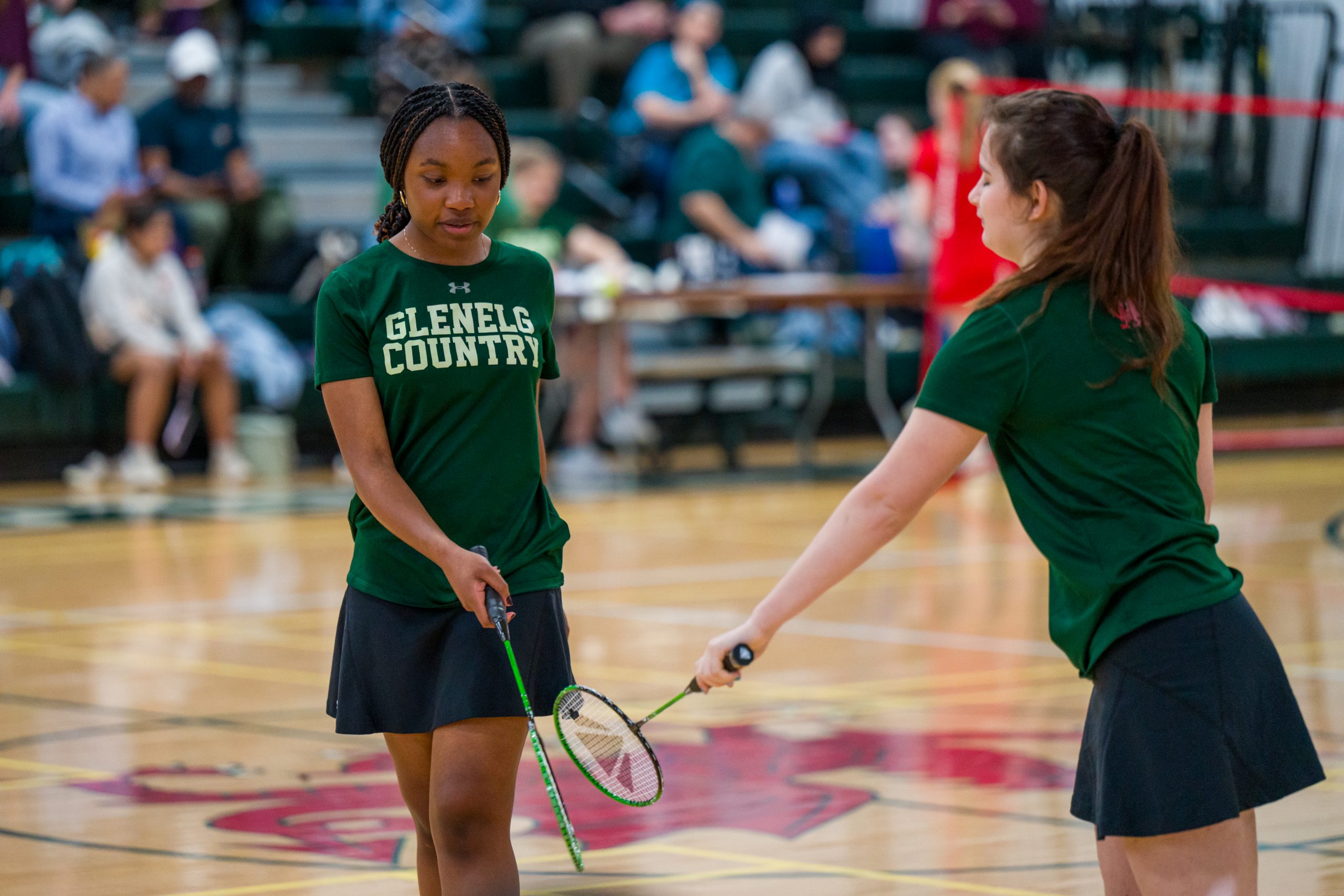 Two female student athletes play badminton. Both girls wear green short sleeve shirt with black skirts. Both hold badminton rackets.