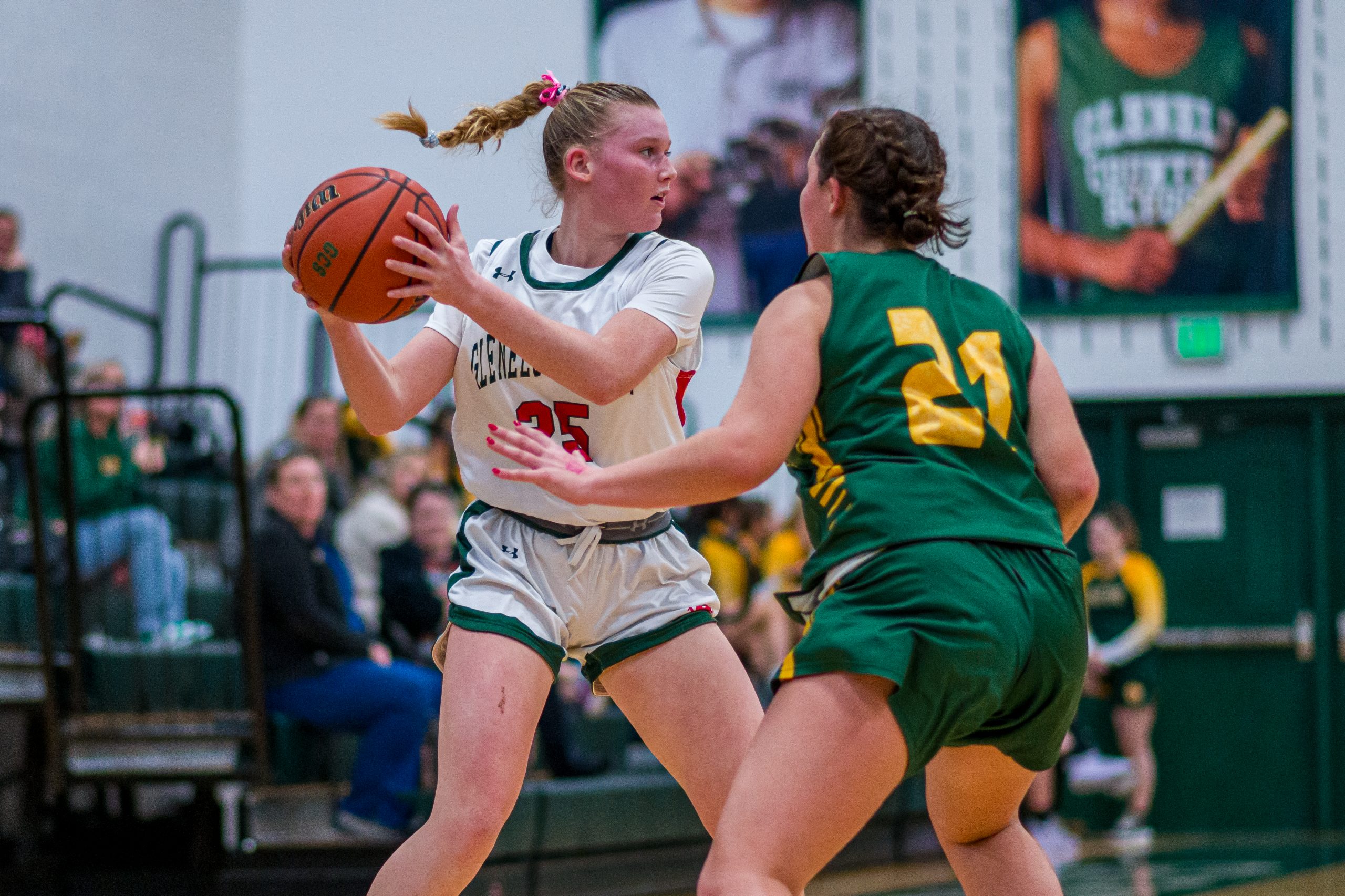 Two female student athletes play basketball. One girl wears a white basketball uniform, and one girl wears a green basketball uniform. One girl holds a basketball.