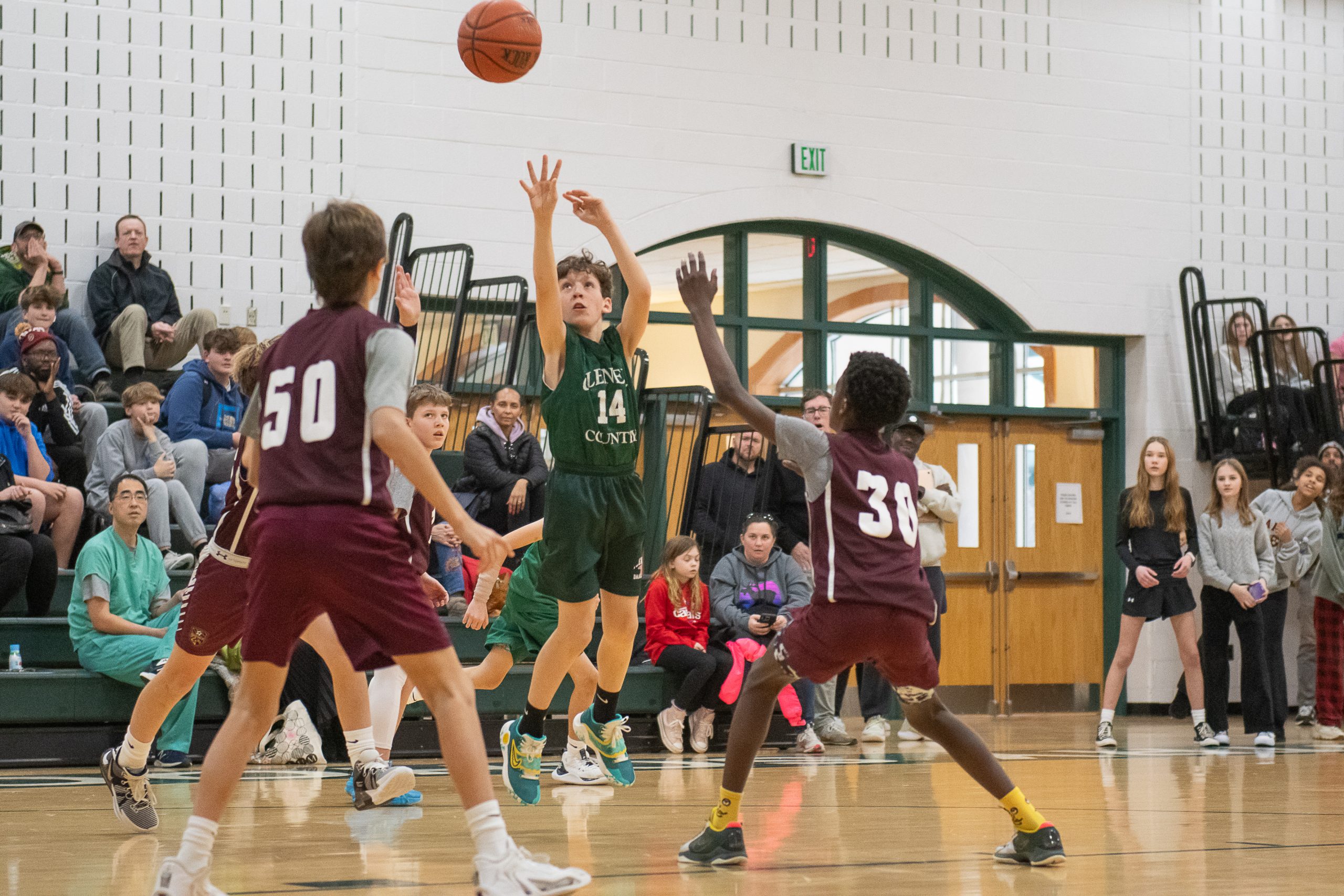 Three male student athletes playing basketball. One student wears a green uniform. Two students wear red uniforms. One student is shooting a basketball.