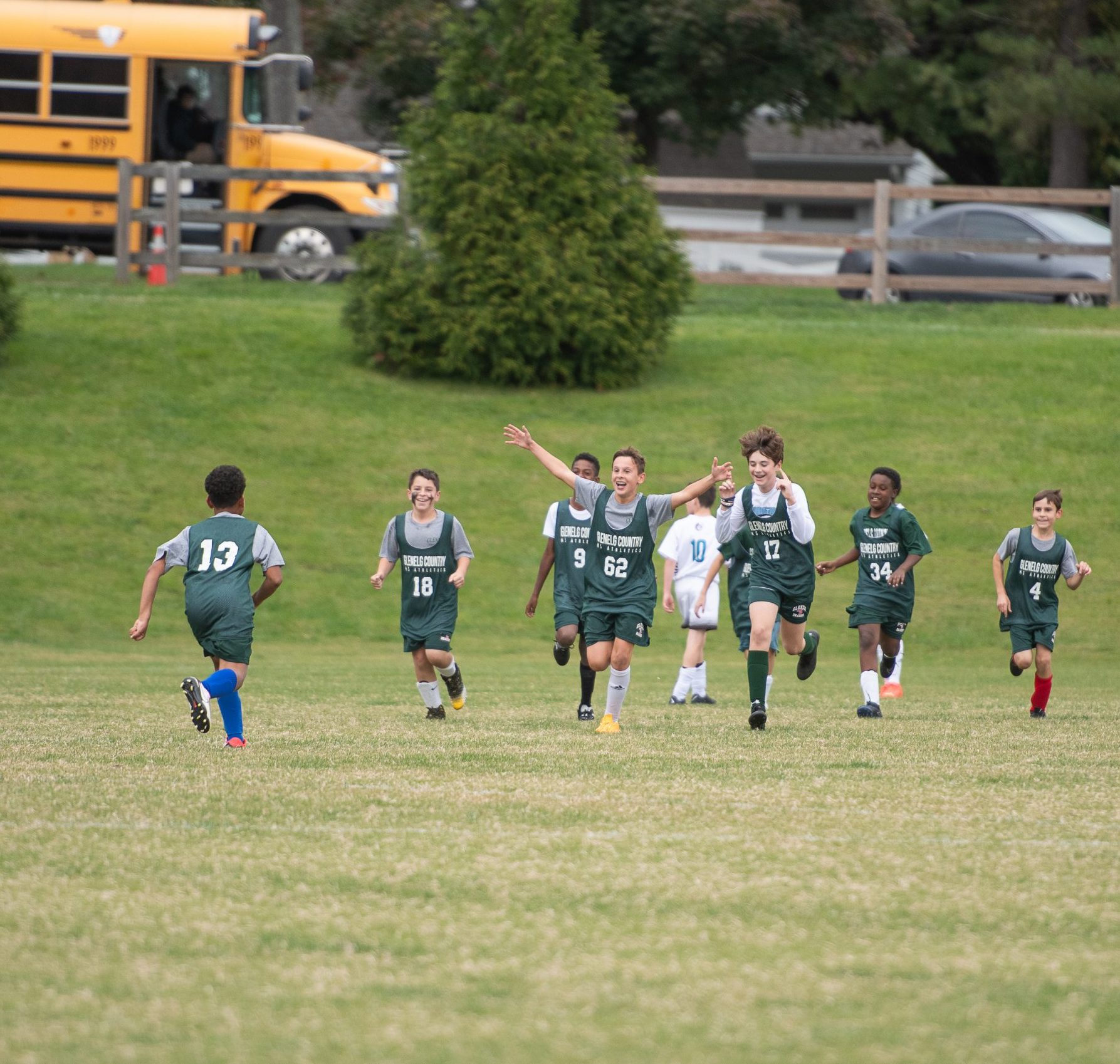 Eight male student athletes playing soccer. Seven students are wearing green uniforms, one is wearing a white uniform. Team celebrates goal.