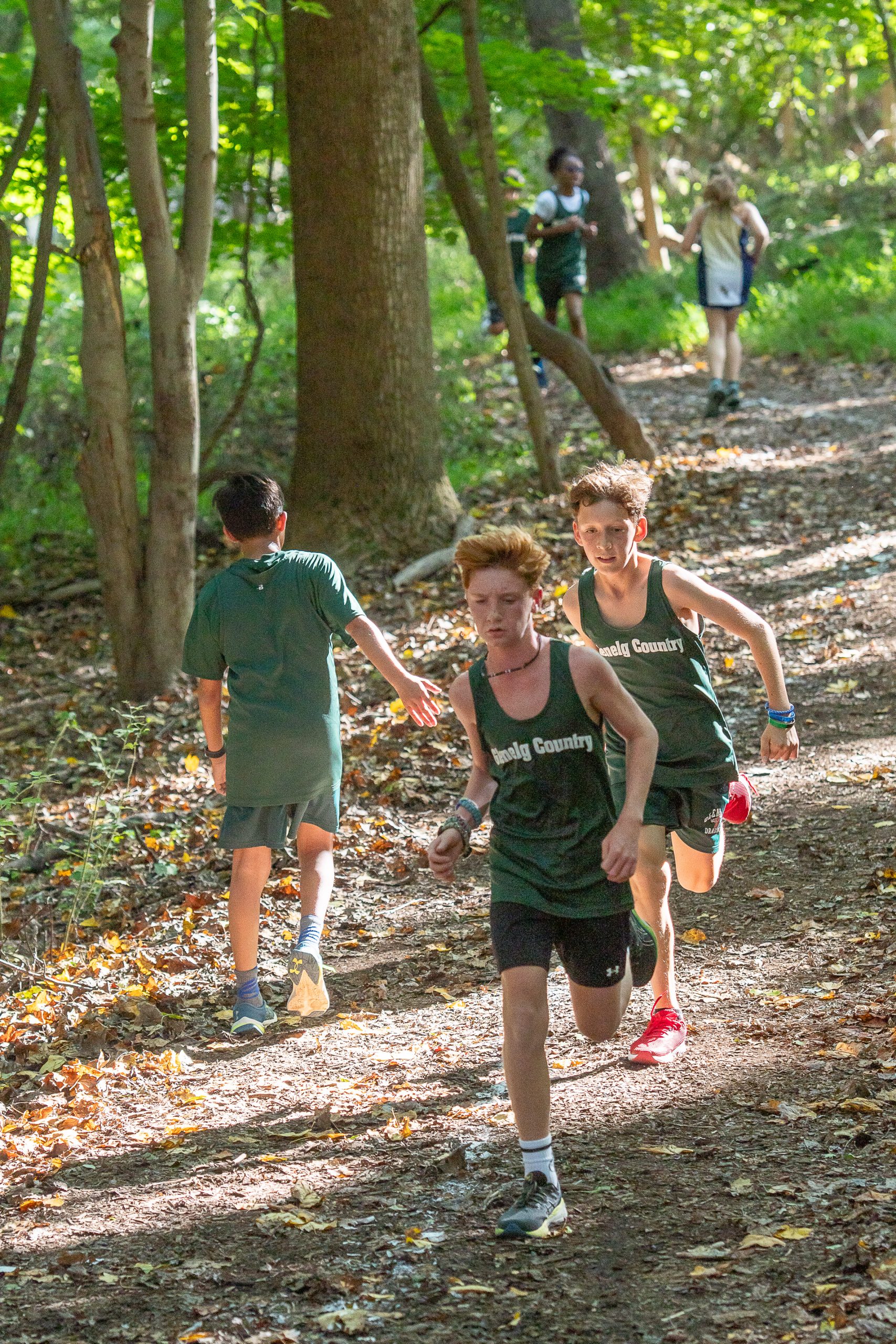 Five student athletes running cross country. Four studetns are wearing green uniforms, one student is wearing a white uniform.