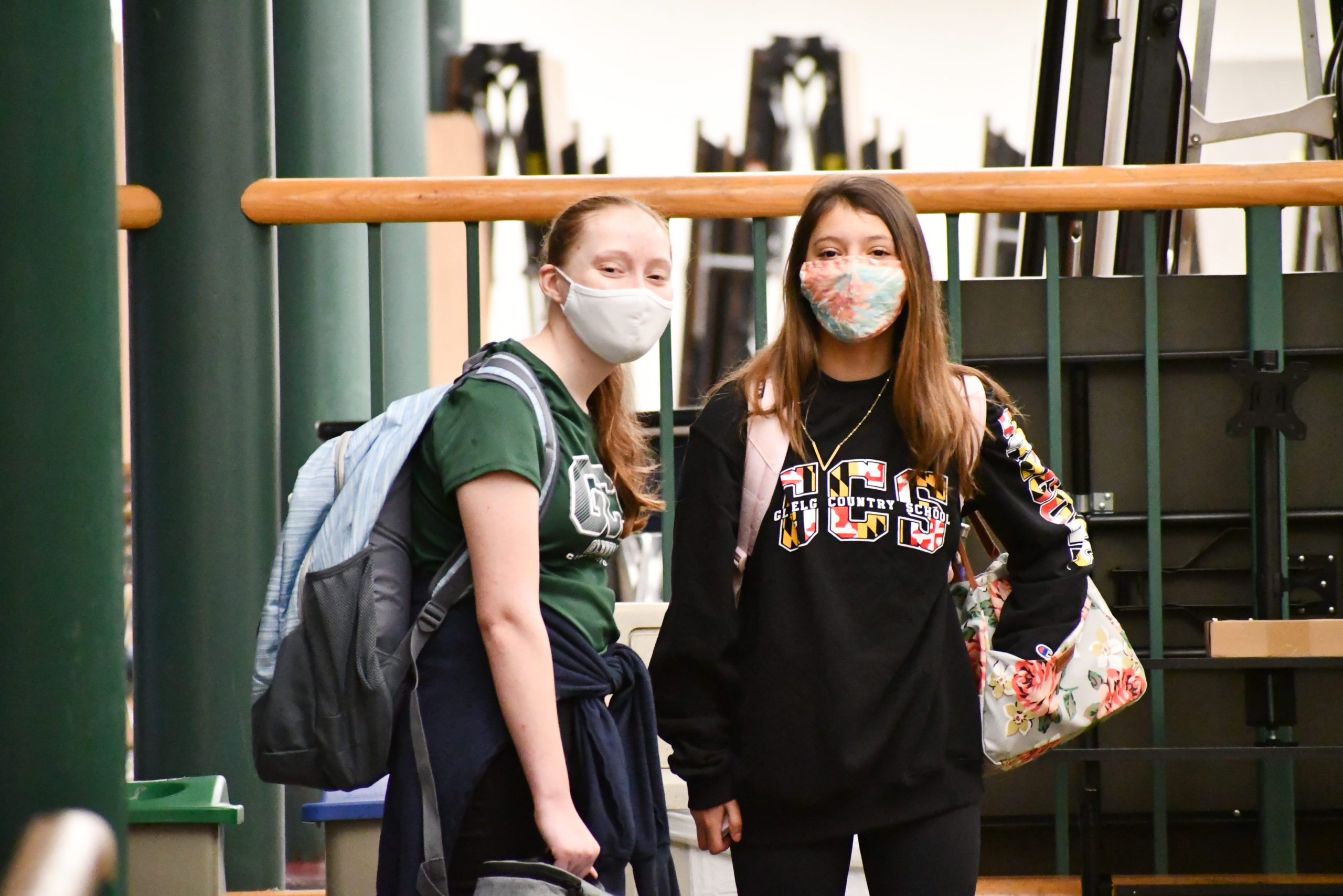 Two female students wearing masks stand in front of a railing inside the school.
