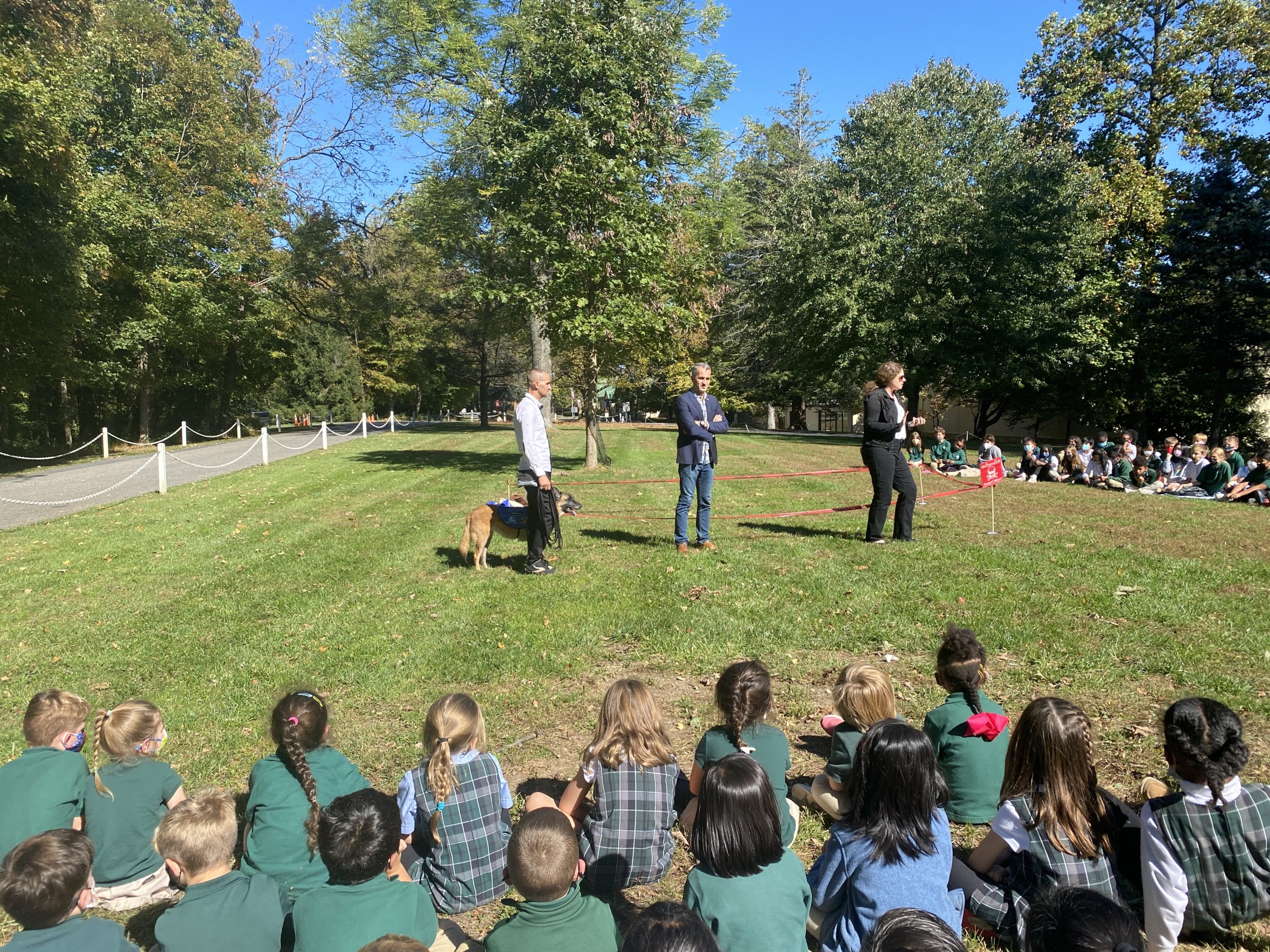 Staff from the Marshall Legacy Institute demostrate a mine detection dog's skills in front of students sitting in a semi-circle on grass.