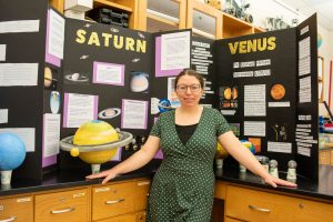 Krystal Rolon, chosen for a space travel educators program, stands in her science lab in front of space diagram featuring the planets Saturn and Venus.