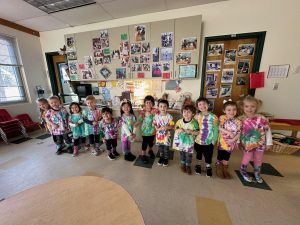 Young students stand in a line insdie a classroom wearing tye-dye shirts. The shirts are mostly white with some color and all the students are wearing black pants or shorts.