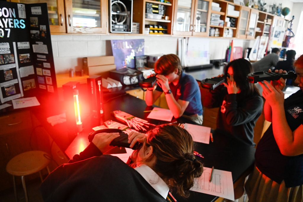 Students in a science lab study spectroscopy as they hold spectroscopes to their face as they look at a red glowing light on the table.