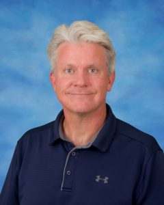 Charlie Stewarts wears a dark navy polo shirt in front of a light blue backdrop as he smiles directly towards the camera.