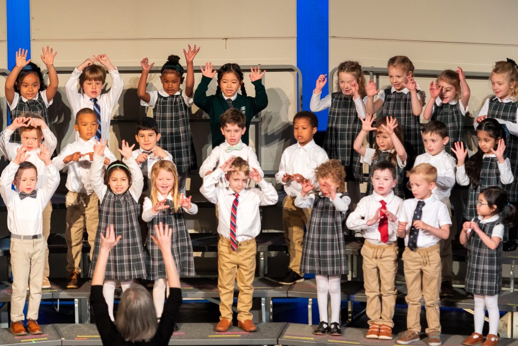 A large group of Kindergarten students put their hands in the air as they perform a song onstage.