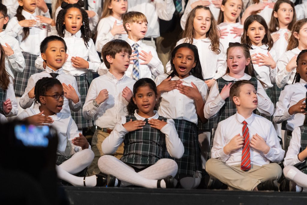 Elementary school children sit on stage and perform a song.