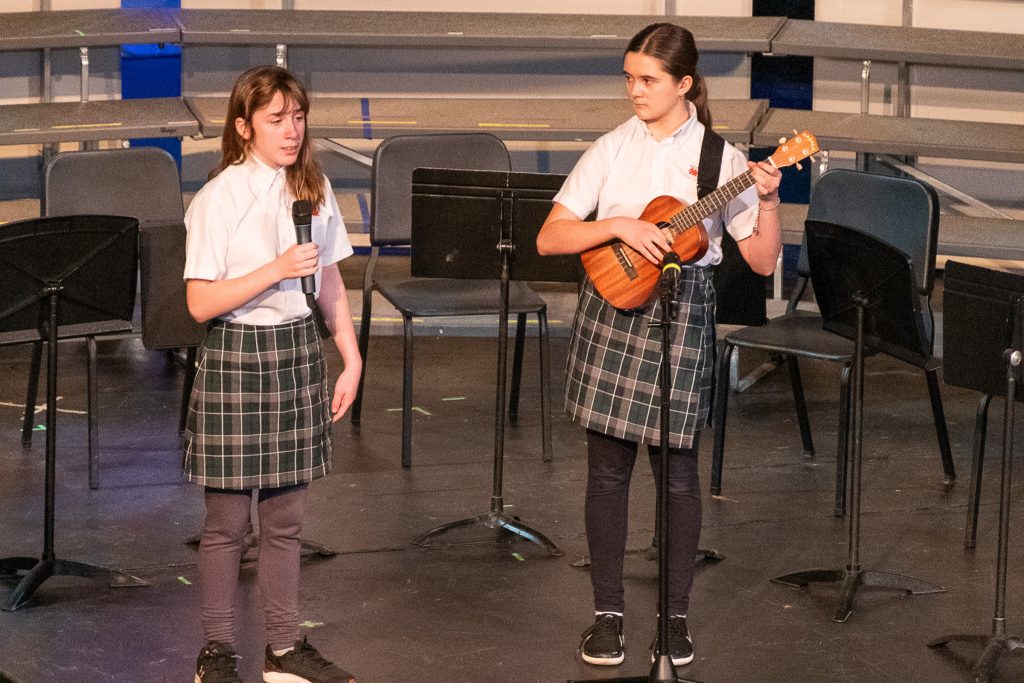 Two middle school girls, one holding a mic and one playing ukelele, perform onstage.