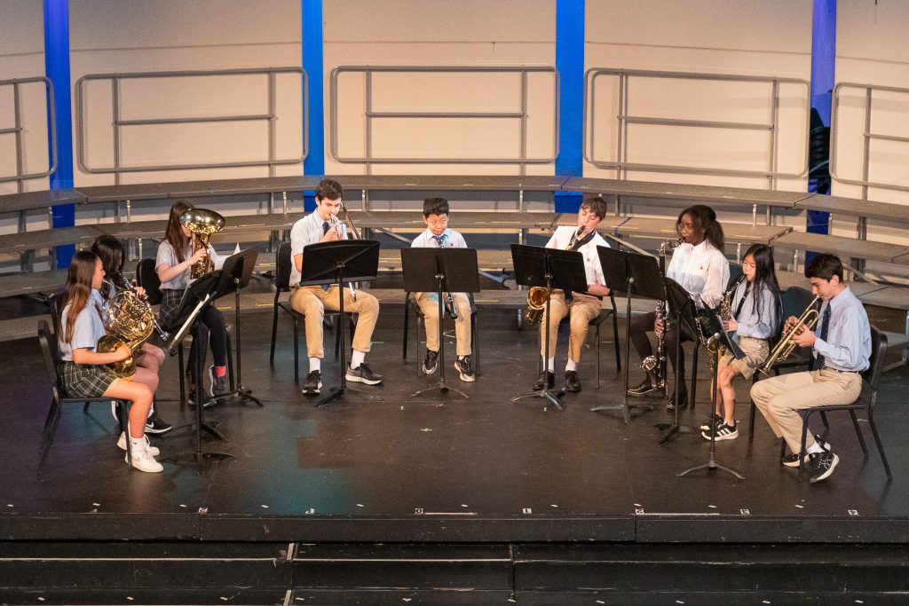Middle School children playing brass and woodwind instruments are seated in semicircle onstage.