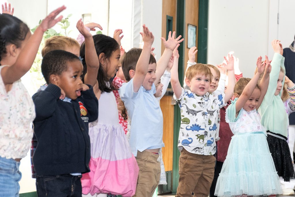 A group of preschool children have their hands in the air as they perform a song together.