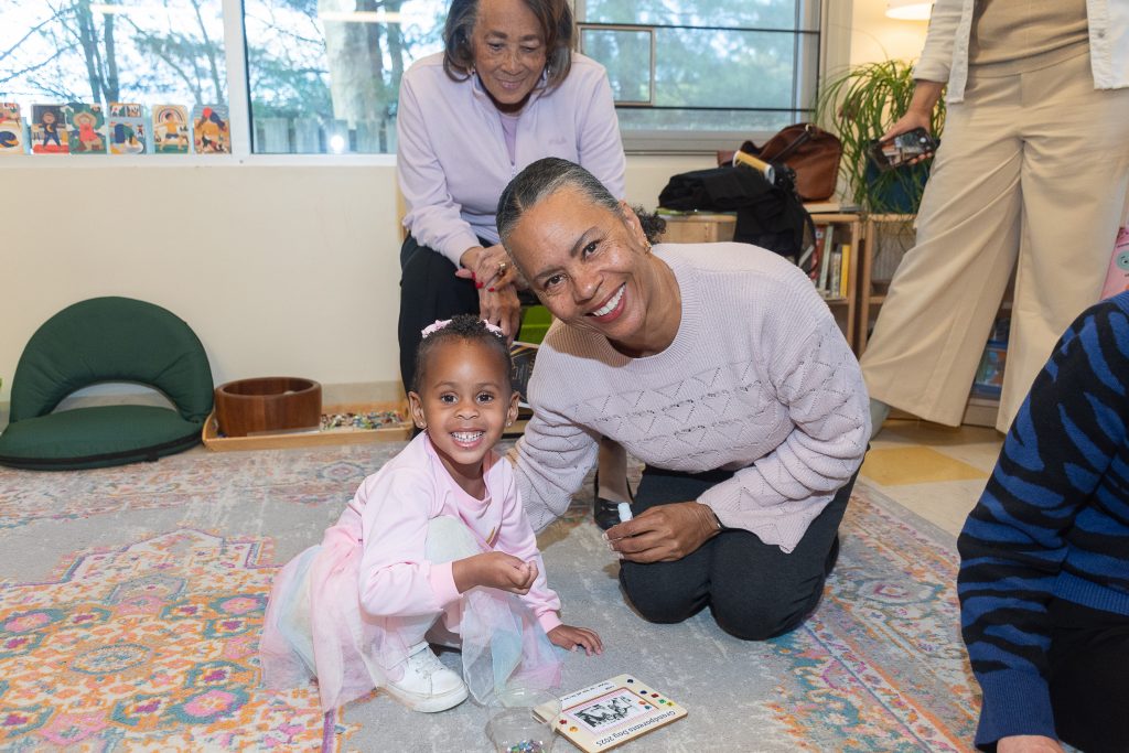 A preschool child and her grandmother pose for a photo in a classroom while another grandmother looks on.
