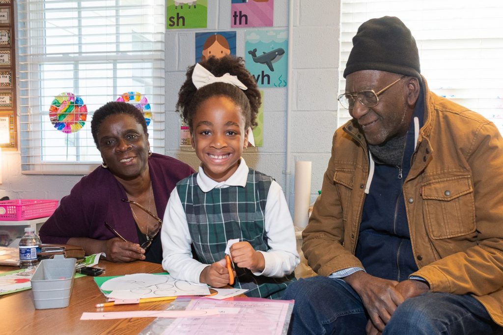 Grandparents sit at a class table with their grandaughter. She is smiling at the camera.